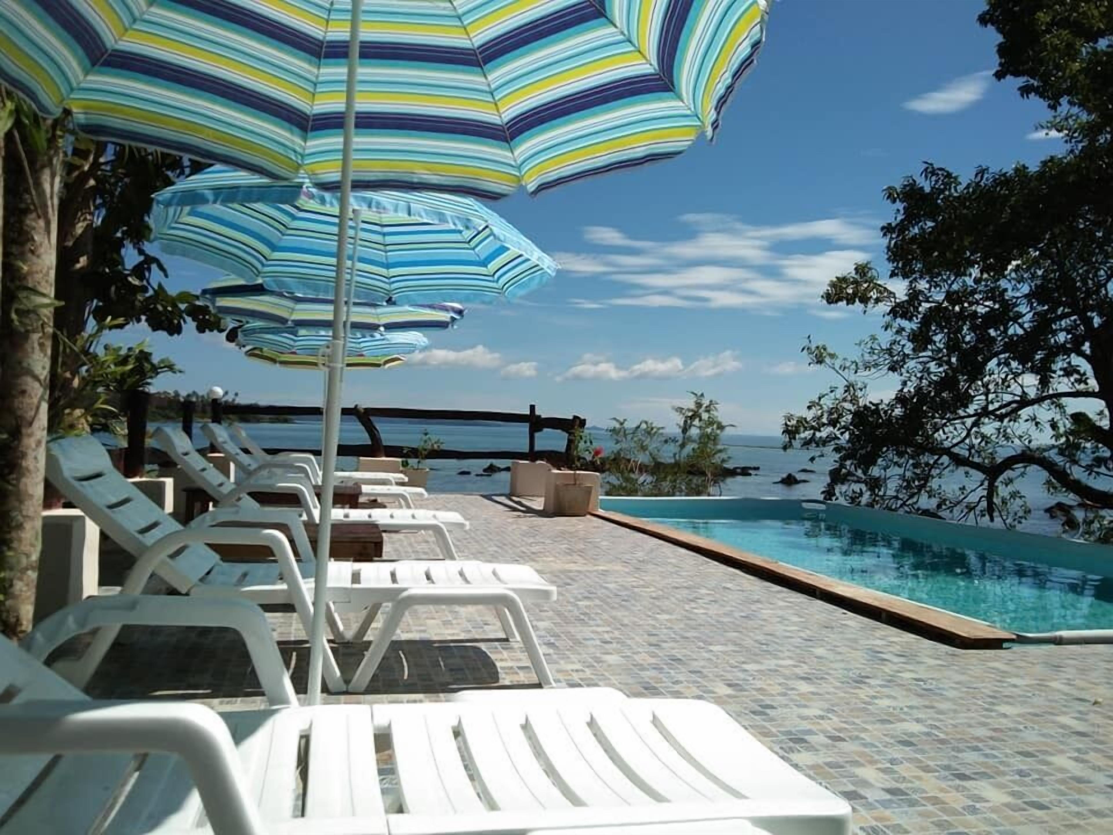 Un espace aménagé au bord de la piscine, avec des chaises longues blanches et des parasols à rayures, offrant une vue sur un ciel bleu dégagé et une mer calme.