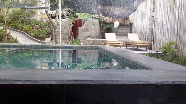 A pool area with lounge chairs, a thatched roof, and a stone wall. - Villa Bintang (Gili Trawangan)