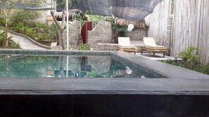A pool area with lounge chairs, a thatched roof, and a stone wall. - Villa Bintang (Gili Trawangan)