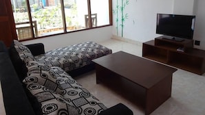 A living room with a sofa, a coffee table, a TV on a wooden stand, and a large window with a view of a garden. - Villa Bintang (Gili Trawangan)