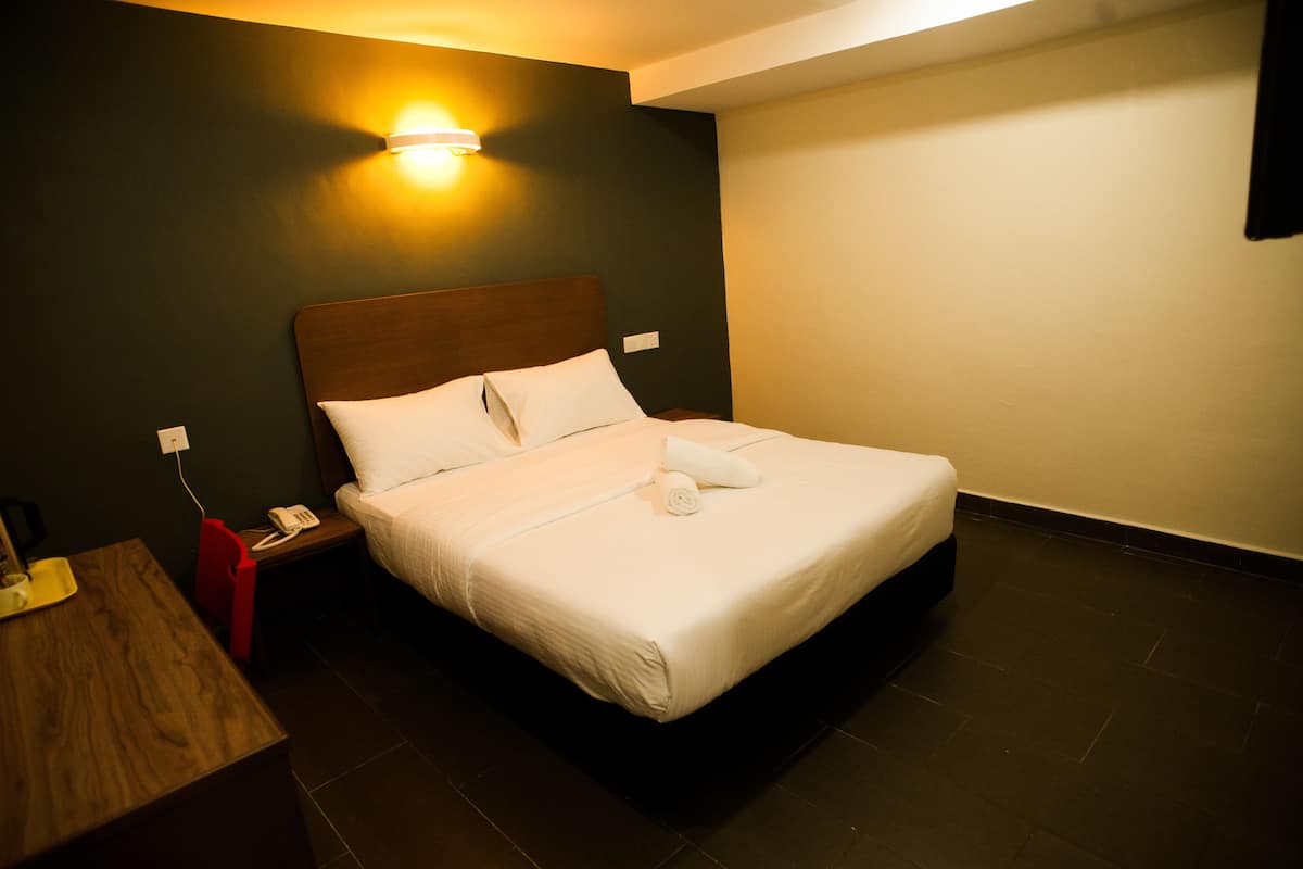 A neatly made bed with white linens and a rolled towel, a wooden nightstand with a phone, and a wall-mounted light fixture.