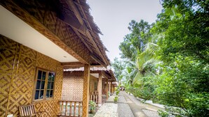 A wooden building with a thatched roof, a covered outdoor seating area, and a pathway leading through a garden with various plants and trees. - Alona Pawikan (Panglao)