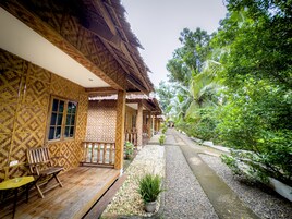Un bâtiment en bois avec un toit de chaume, un espace extérieur abrité avec des sièges, et un sentier qui traverse un jardin où l’on trouve différentes plantes et arbres.