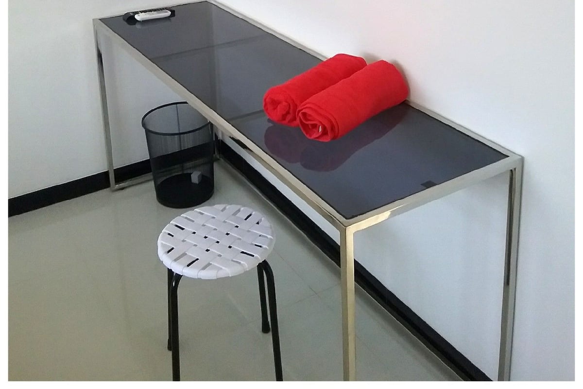 A small, modern room with a glass-top desk, a black TV mounted on the wall, a white chair, and a pair of red towels on the desk.