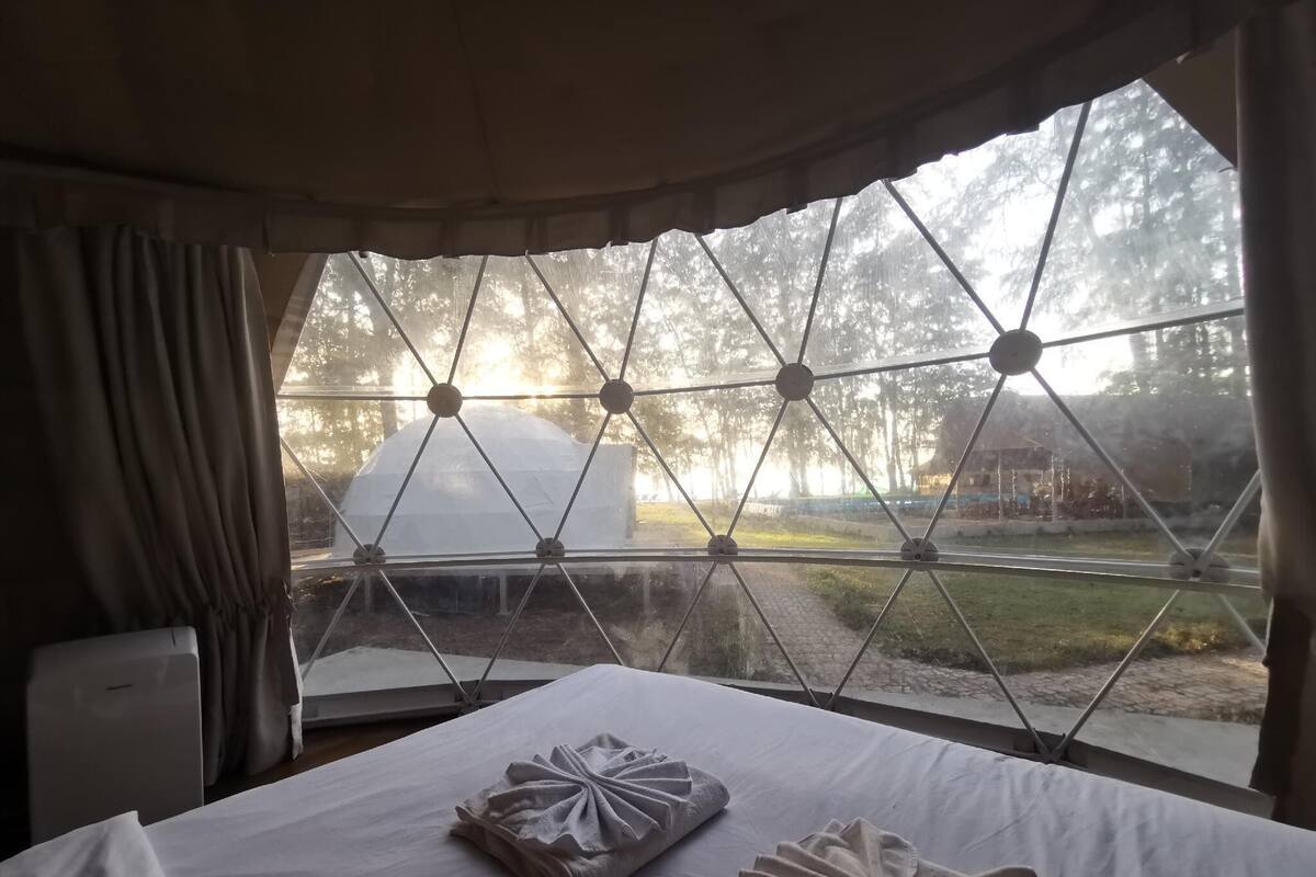 Une chambre avec une grande fenêtre, un lit où les serviettes sont soigneusement pliées, et une vue sur une tente et des arbres à l’extérieur.