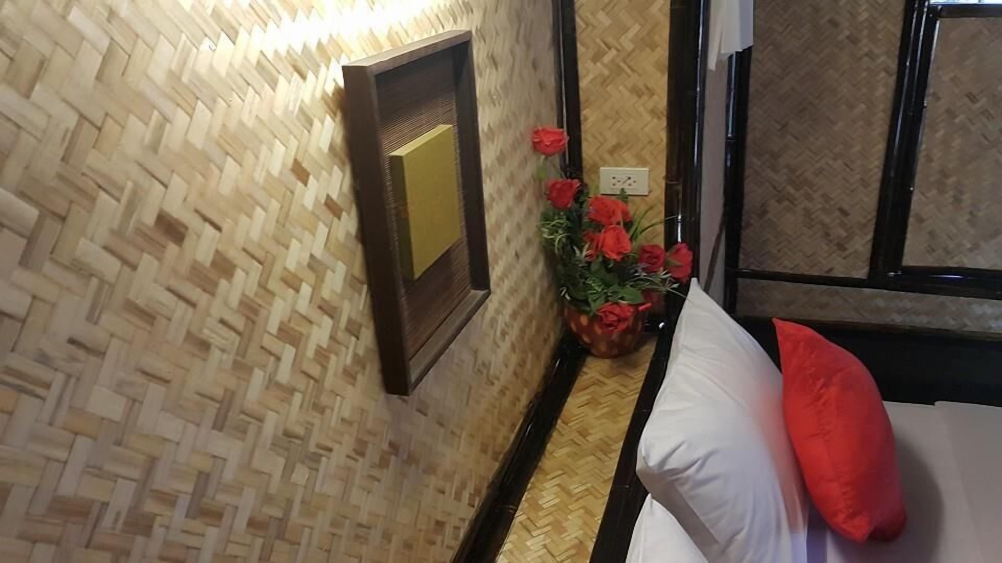 Habitación de hotel con pared tejida, cama con almohada blanca y almohada decorativa roja, y planta en maceta con flores rojas.
