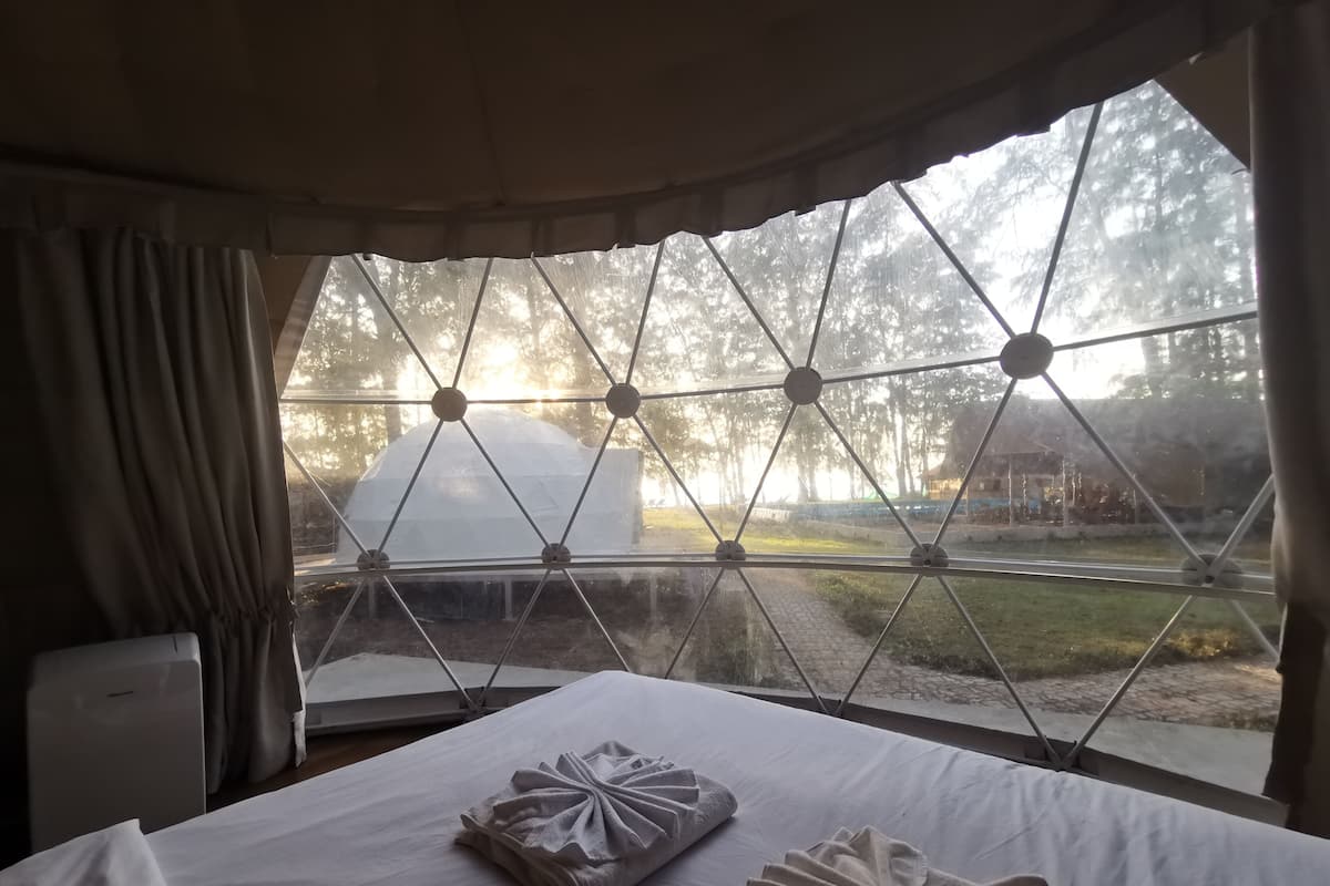 Une chambre avec une grande fenêtre, un lit où les serviettes sont soigneusement pliées, et une vue sur une tente et des arbres à l’extérieur.