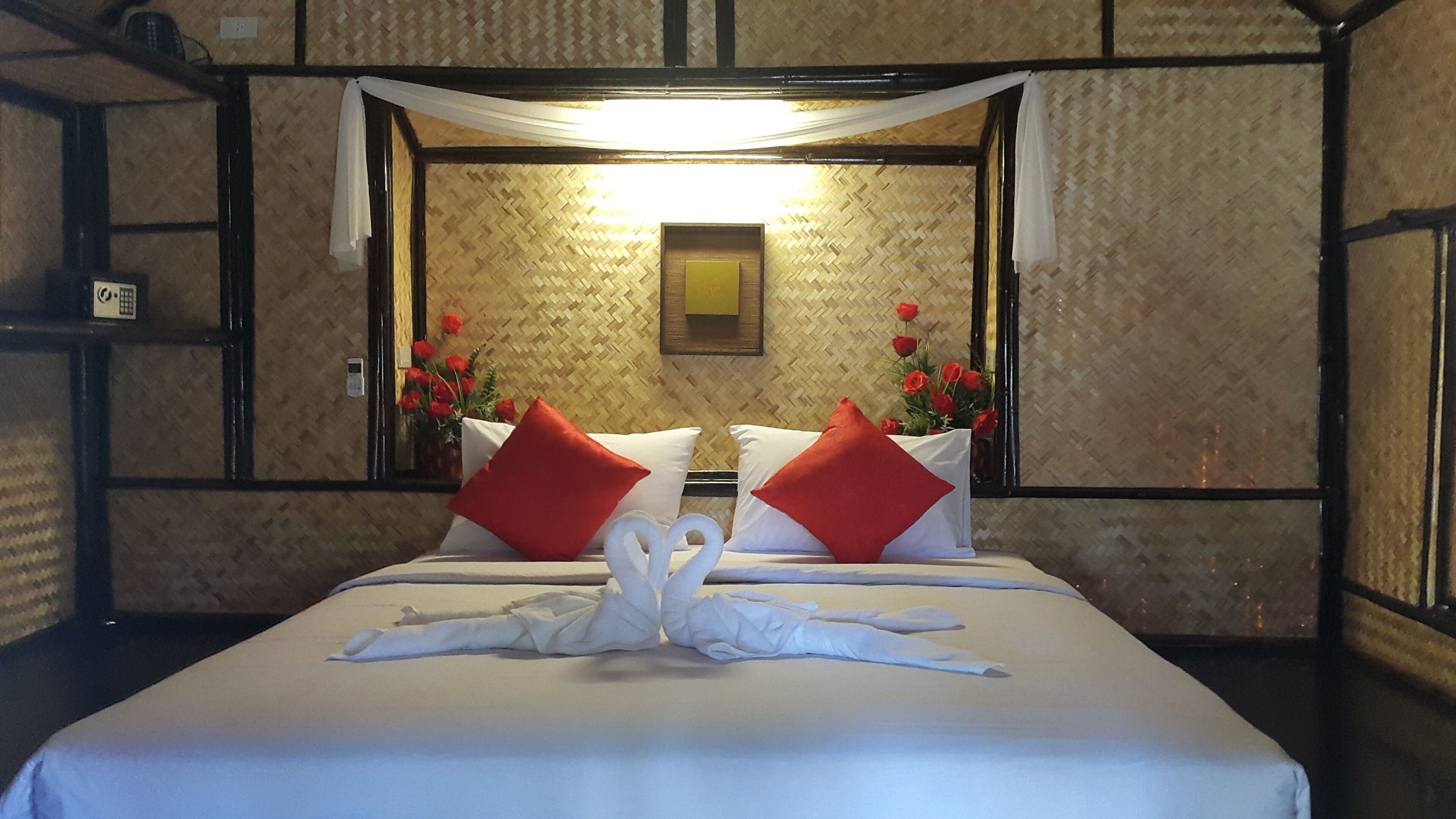 Een hotelkamer met een bed, wit beddengoed, rode kussens, een witte handdoekstapel en een wandlamp, plus een lijst met een afbeelding.