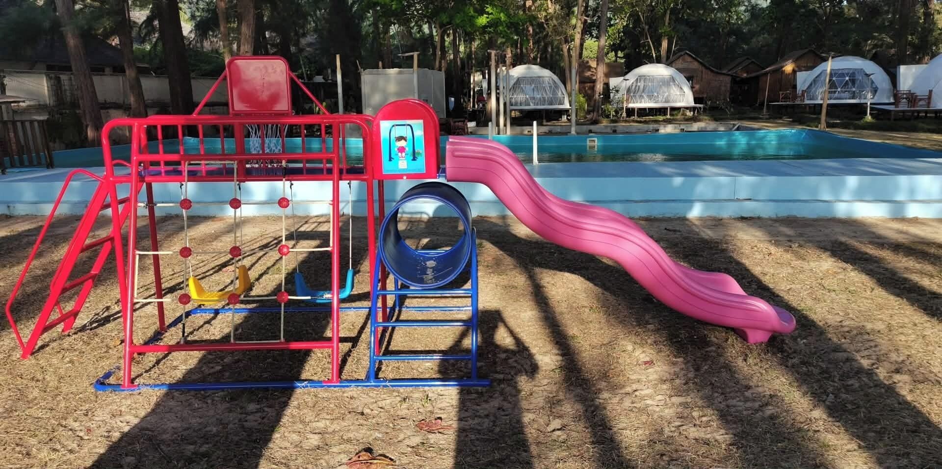 Un parque infantil con tobogán y columpios, rodeado de árboles y una piscina.
