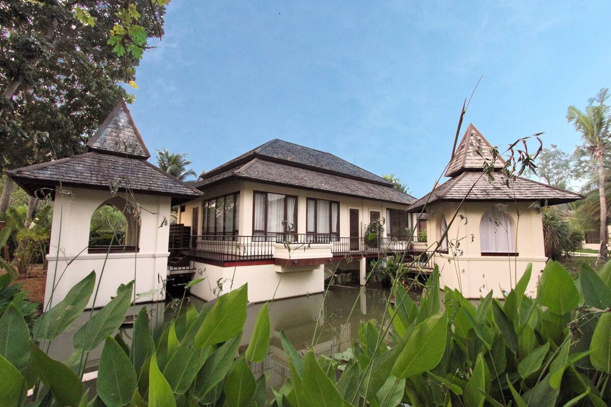 A traditional house with a central building and two smaller structures with pointed roofs, surrounded by lush greenery and a calm water body.
