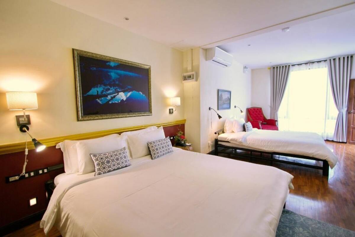 A hotel room with two beds, a large painting, a red chair, and a window with curtains.