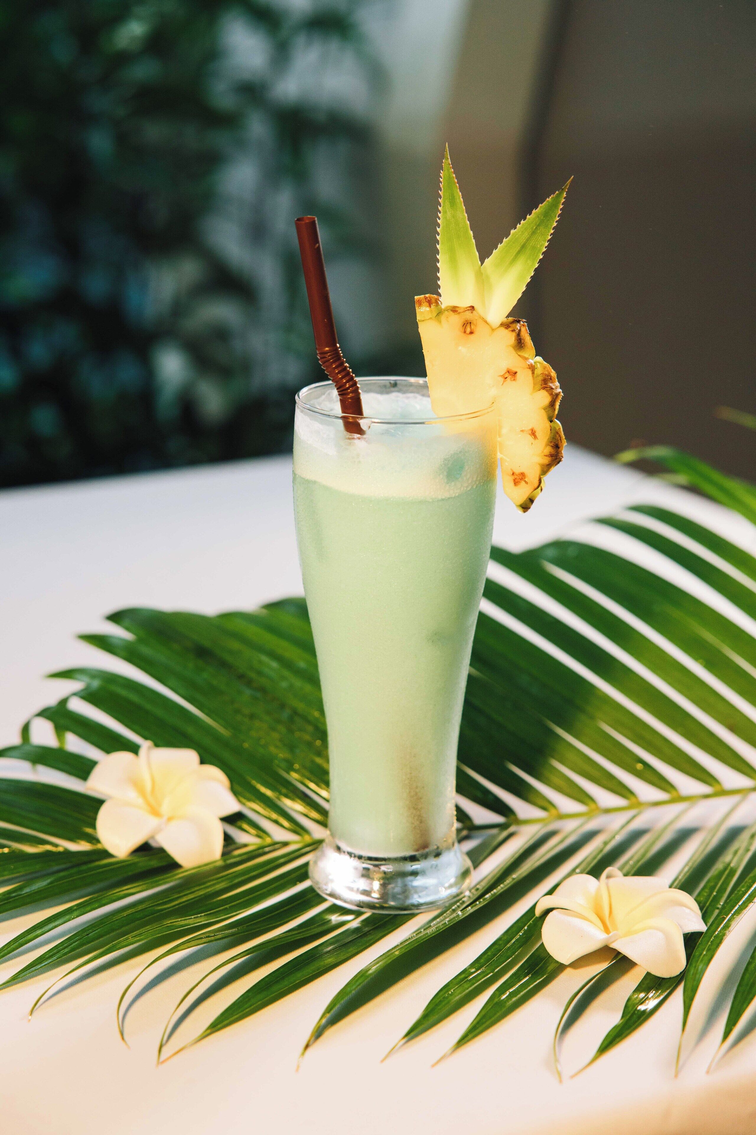 A tall glass with a straw, garnished with a pineapple slice and a flower, placed on a surface with green leaves and a white flower.