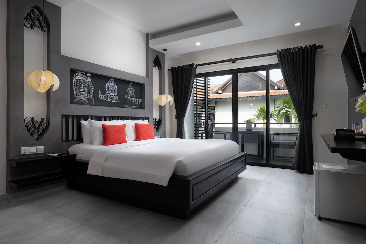 A modern bedroom with a large bed, a flat-screen TV, a small desk, and a balcony with a view of the outside.