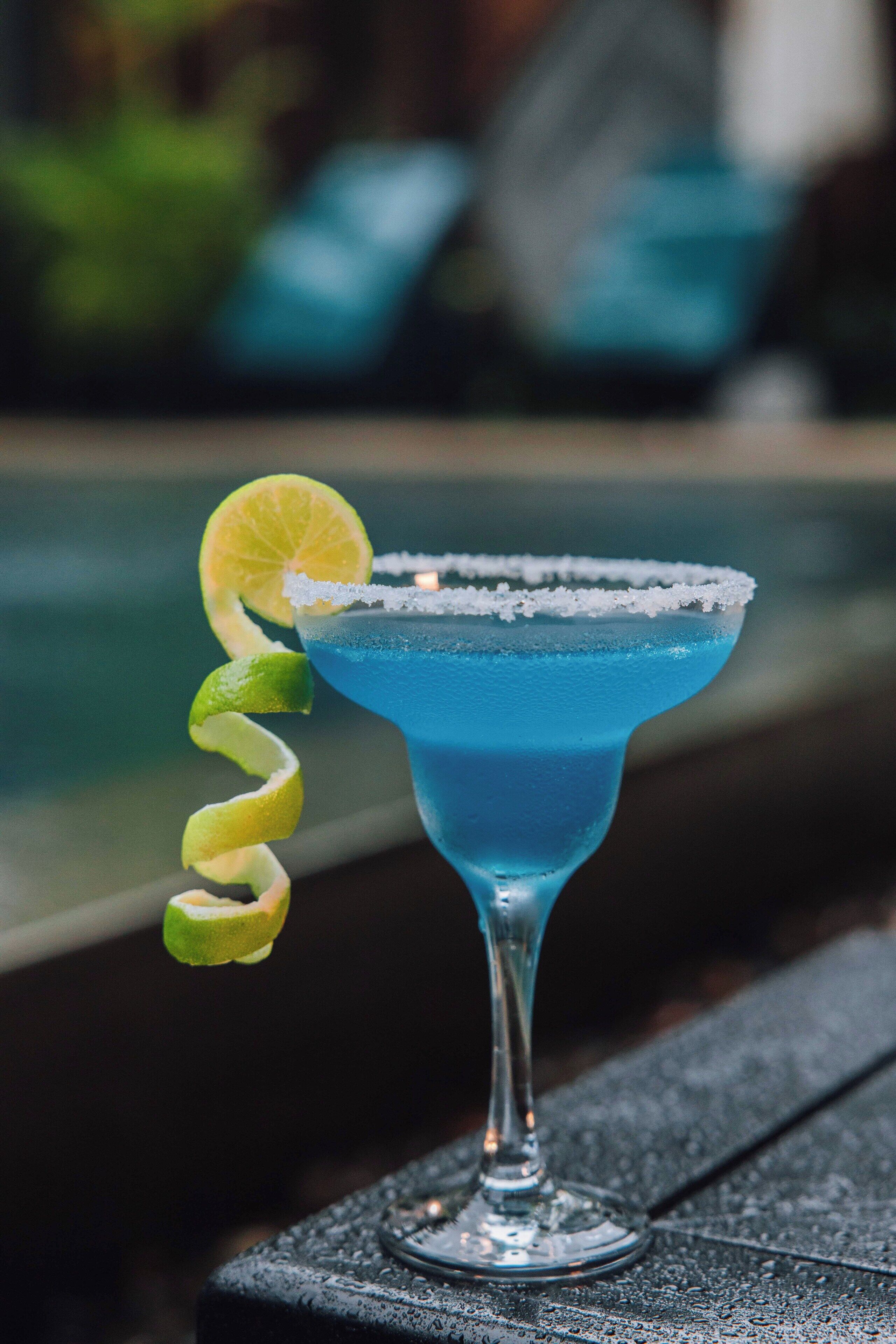A blue cocktail with a lime garnish and salt rim, placed on a wooden surface.