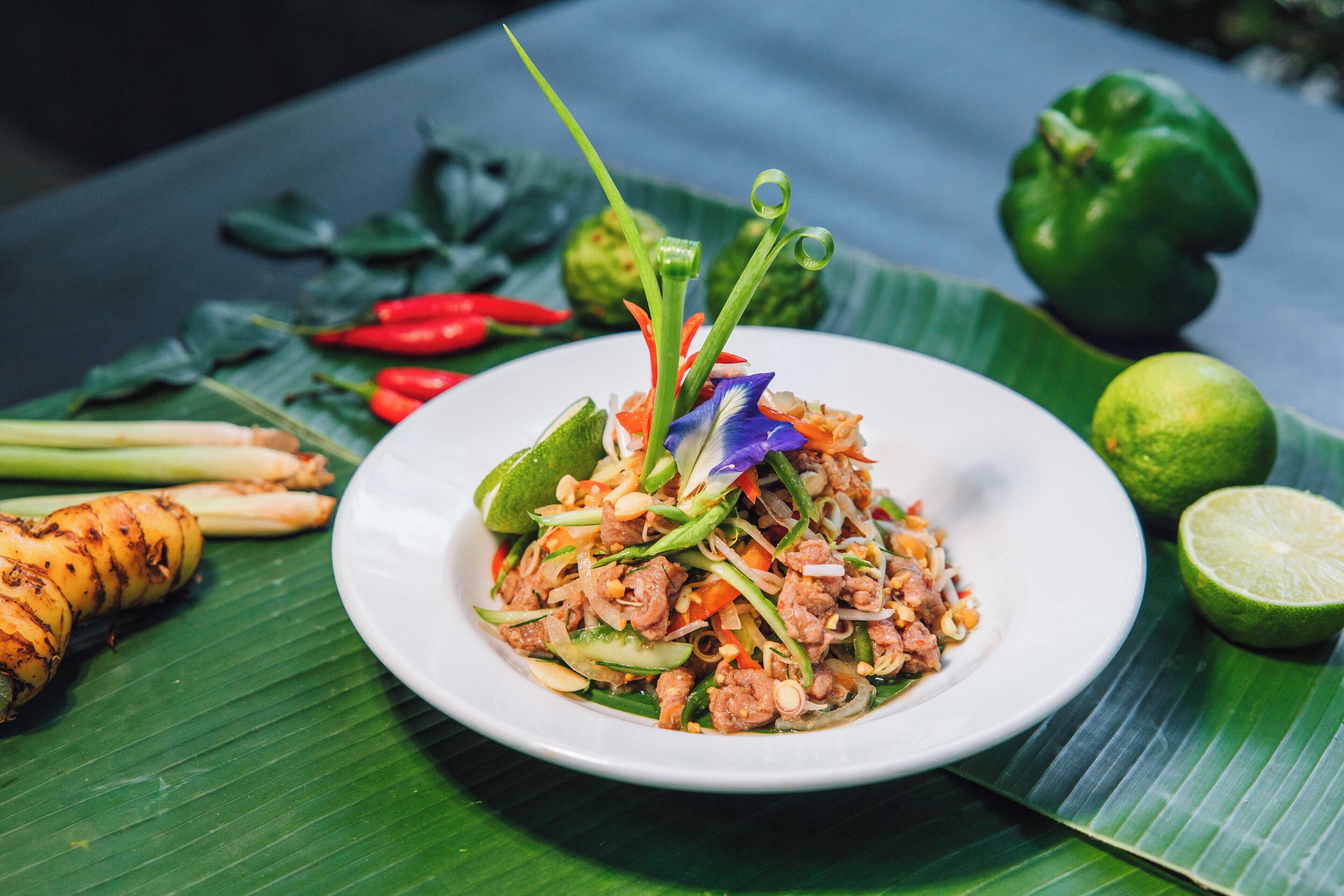 A plate of food with various vegetables and meat, garnished with green onions and a flower, on a white plate with a banana leaf underneath.