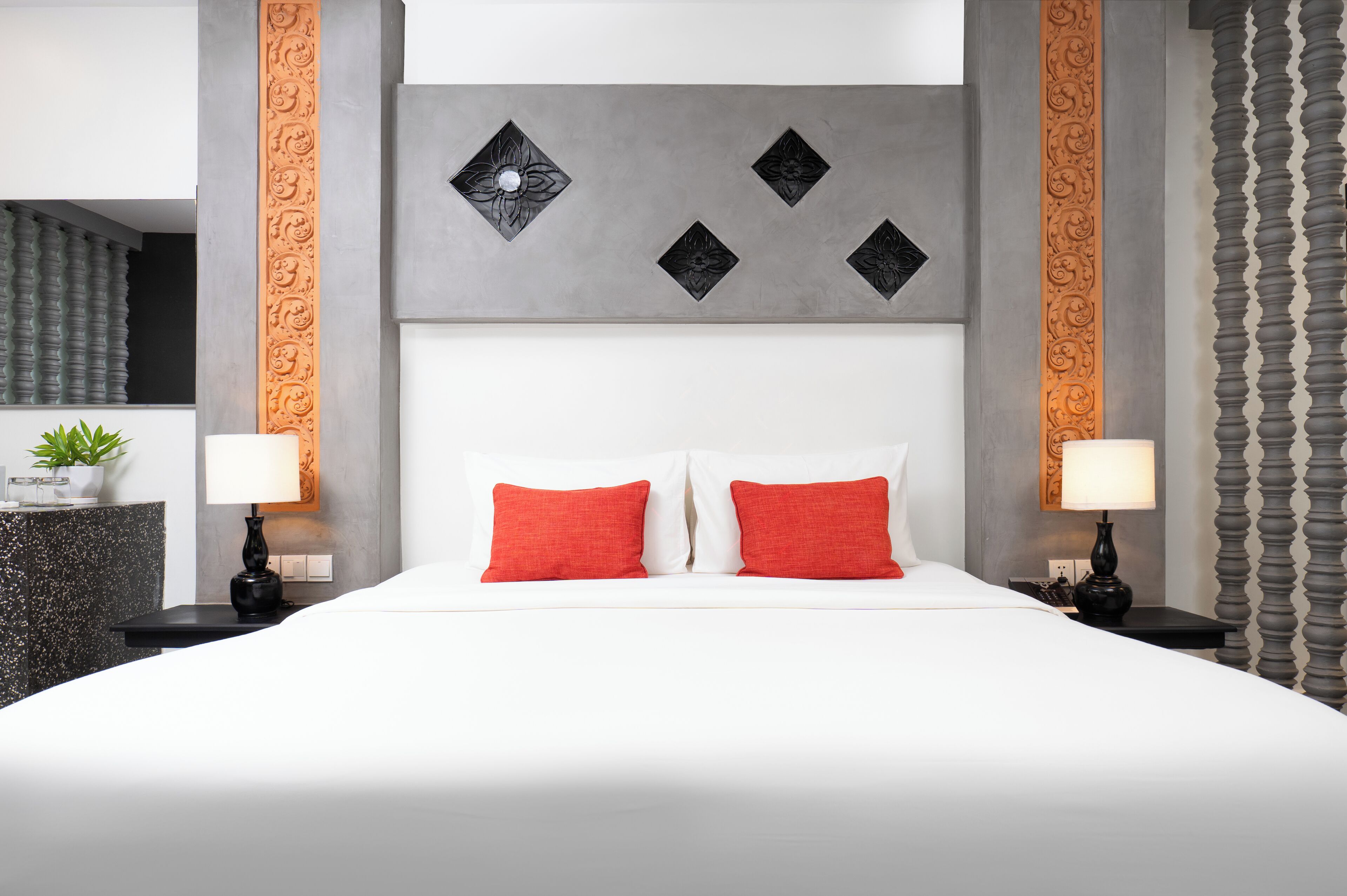 A modern hotel room with a large bed, two red pillows, a grey headboard with decorative panels, and a nightstand with a lamp and a small plant.