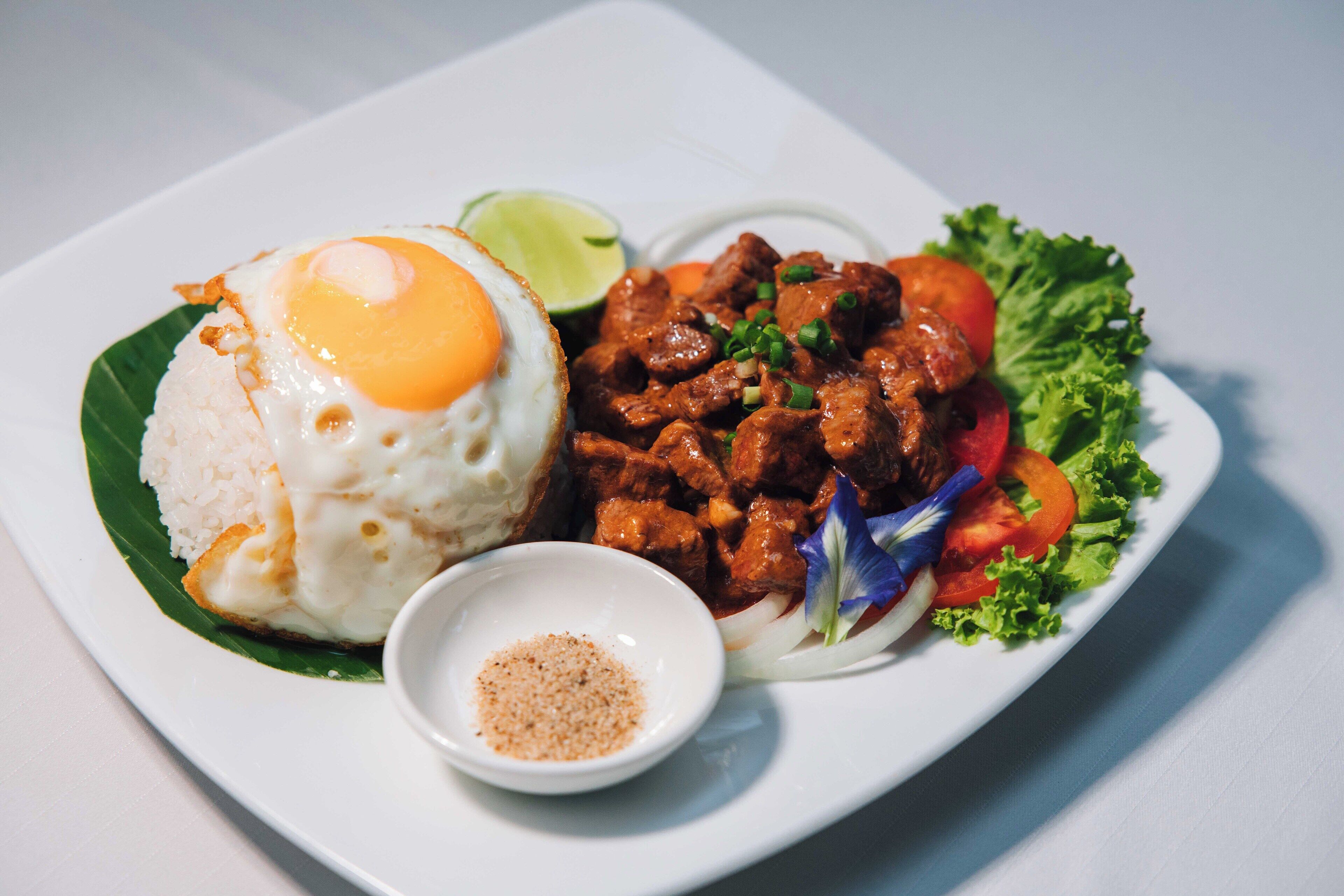 A plate with rice, a fried egg, meat, vegetables, and a small bowl of seasoning.