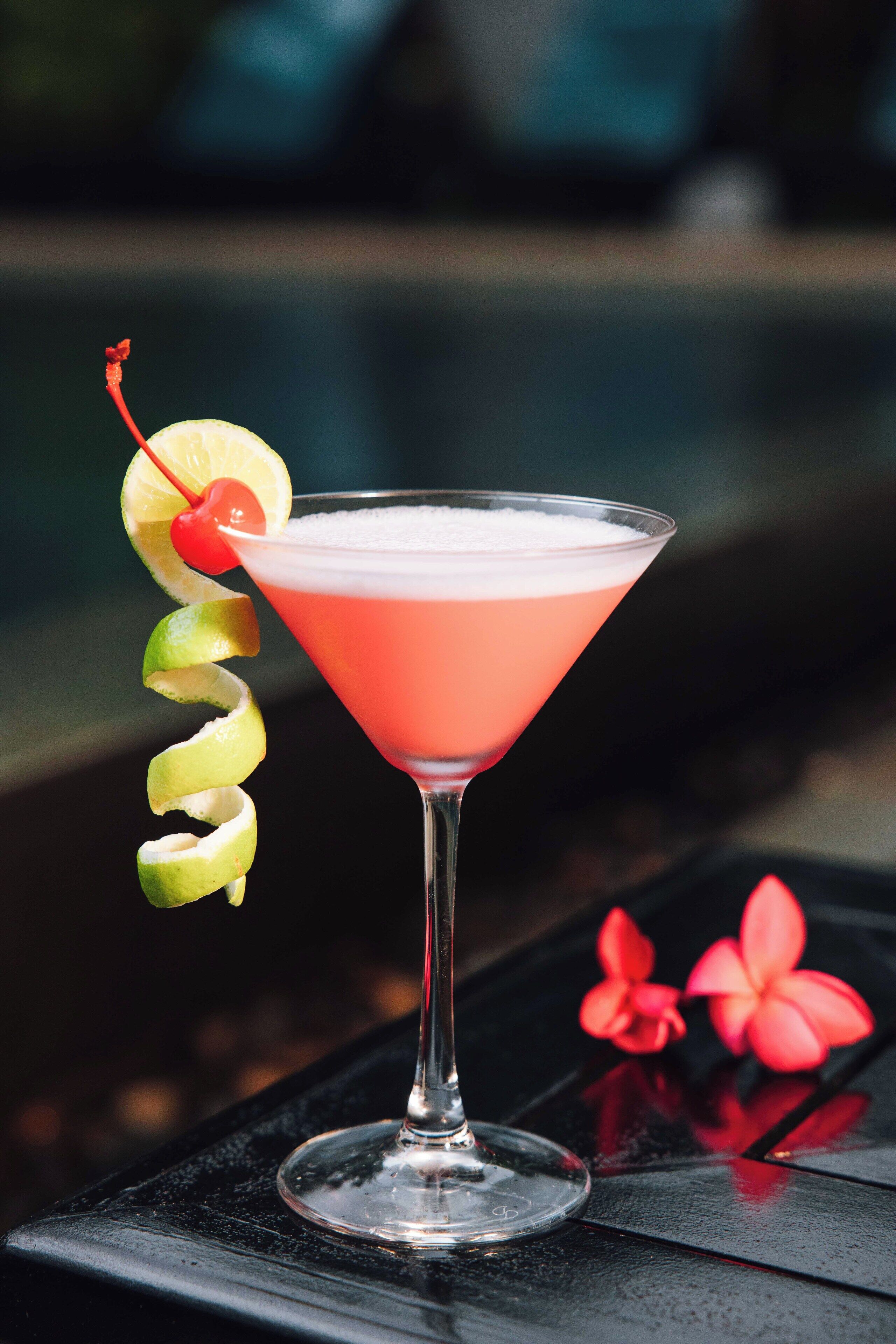 A cocktail in a martini glass with a cherry and lime garnish on a black surface.