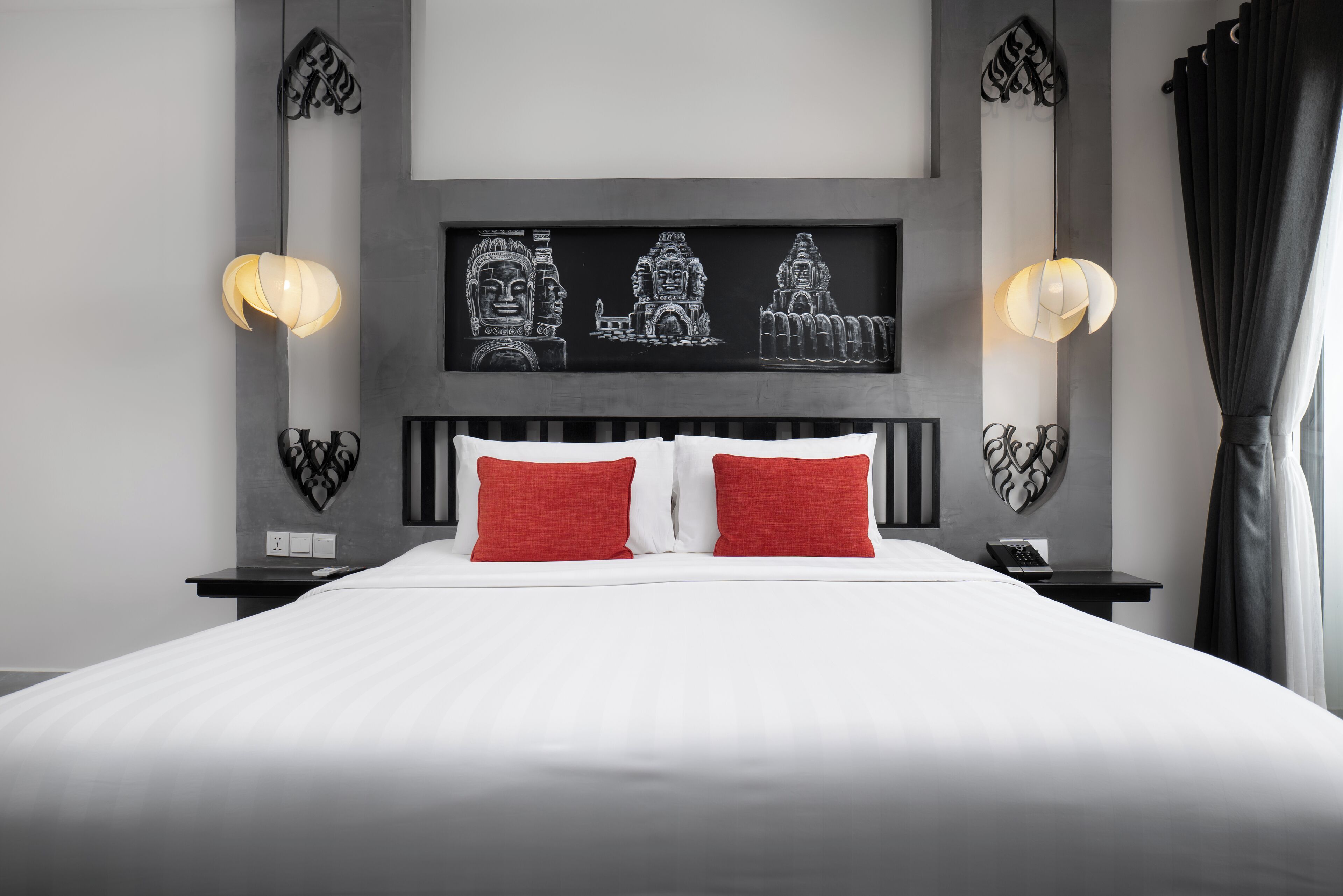A modern hotel room with a large bed, two red pillows, a black headboard, and wall art depicting ancient temples.