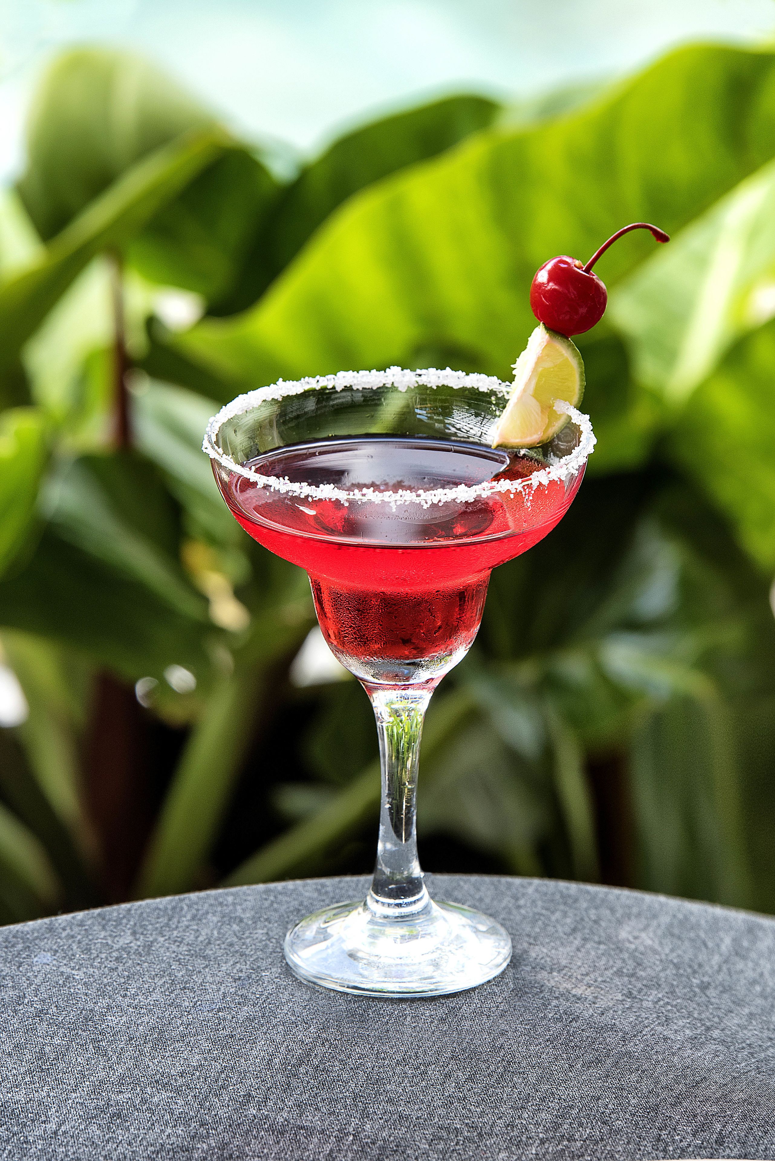 A cocktail in a glass with a cherry garnish, set against a blurred green background.