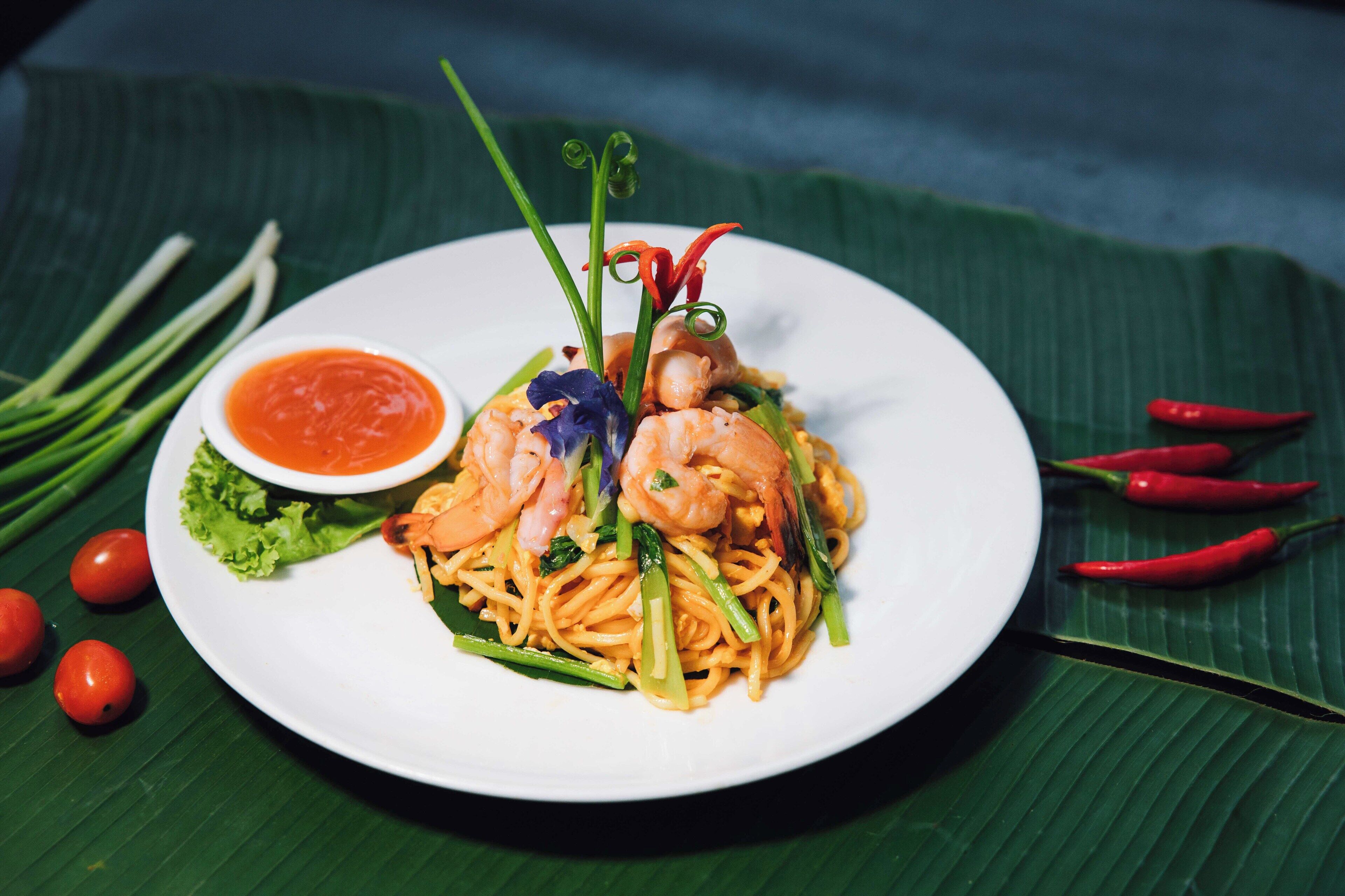 A plate of noodles with shrimp and vegetables, a small bowl of sauce, and chili peppers on a banana leaf.