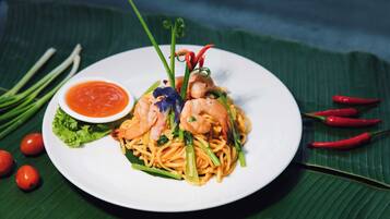 A plate of noodles with shrimp and vegetables, a small bowl of sauce, and chili peppers on a banana leaf.