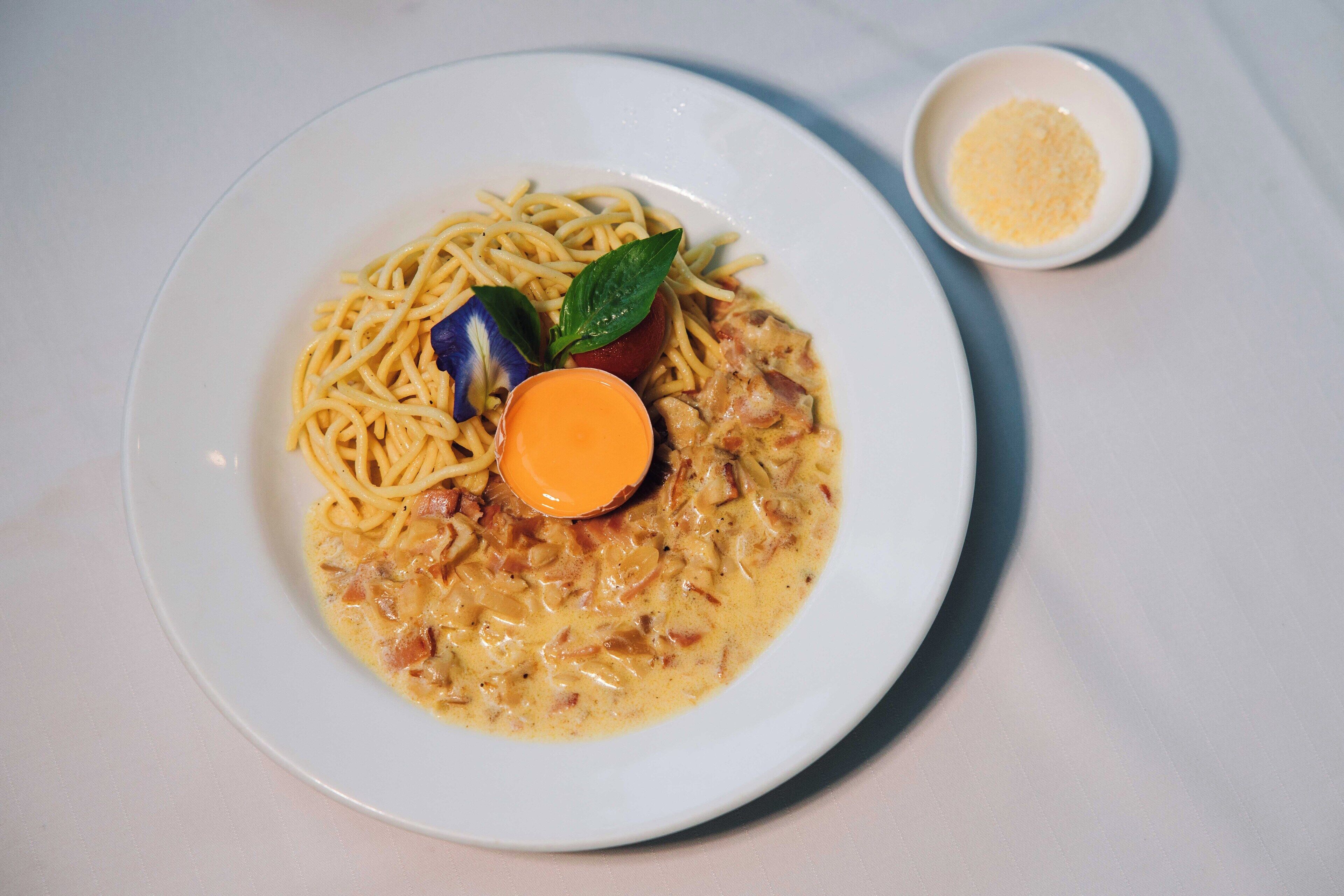 A plate of spaghetti with a creamy sauce and a piece of meat, garnished with a basil leaf and a slice of orange.