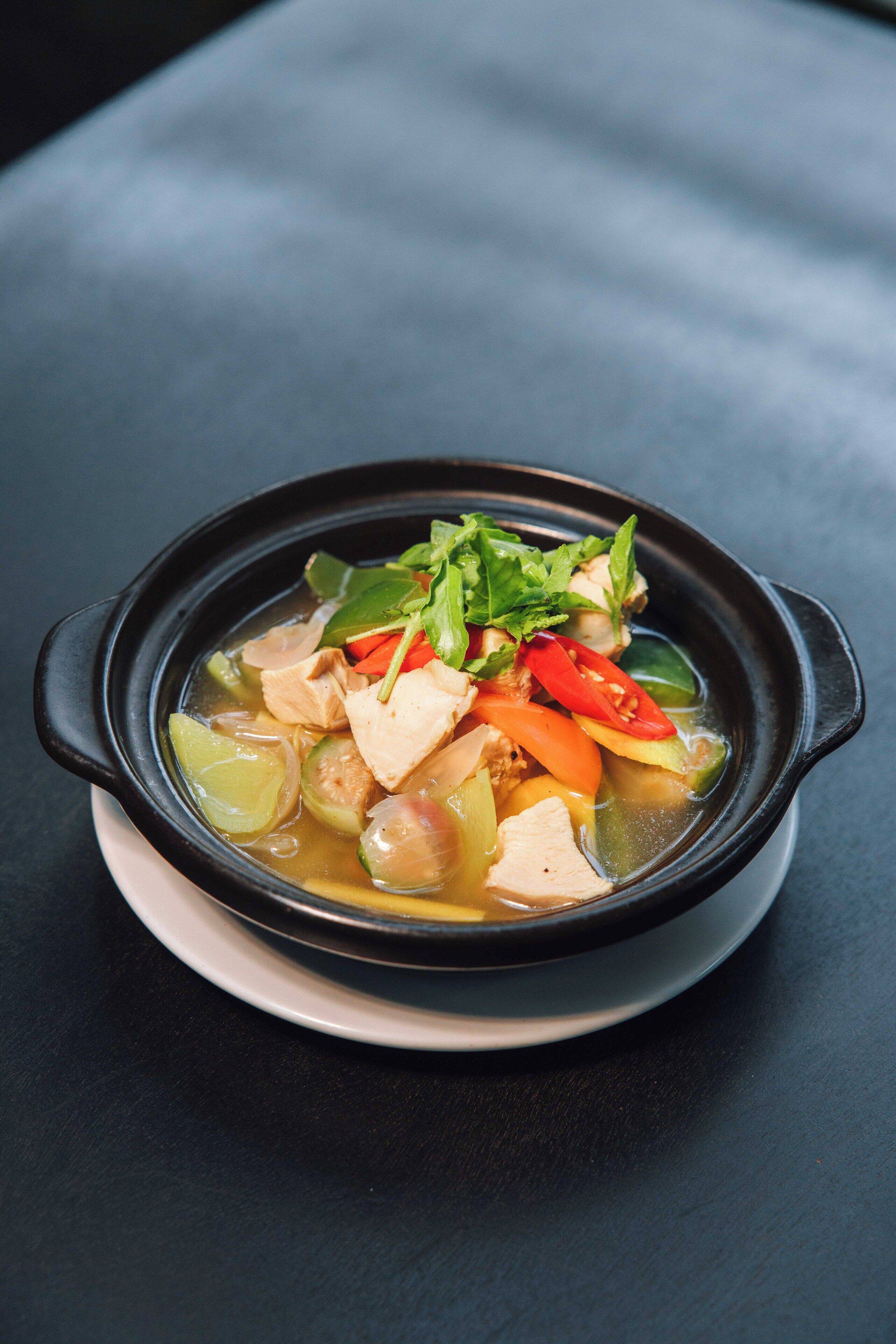 A bowl of soup with various vegetables and meat, served in a black pot on a white plate.