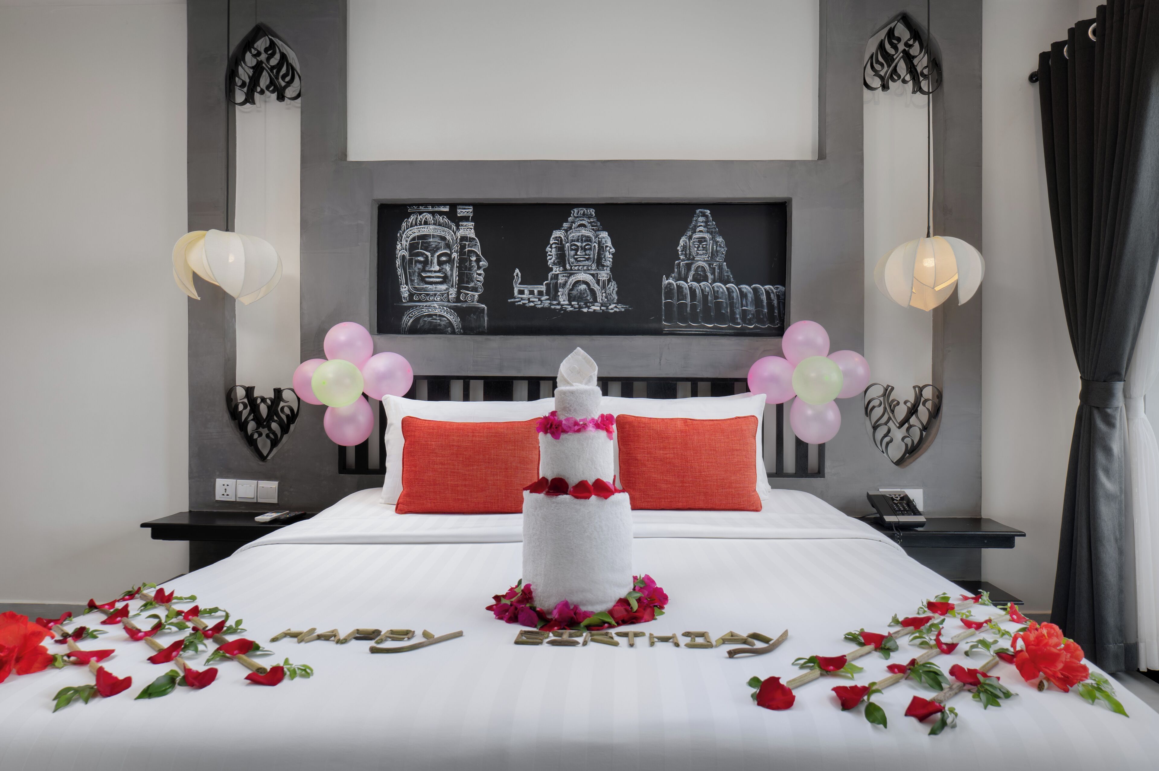 Una habitación de hotel con una cama, una obra de arte en la pared que representa rostros de templos, una mesita de noche con un teléfono y una lámpara.