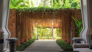 A serene indoor garden with a brick wall, a hanging bamboo curtain, and a pathway leading to a seating area with a sofa and a table. - Central Suite Residence (Siem Reap)
