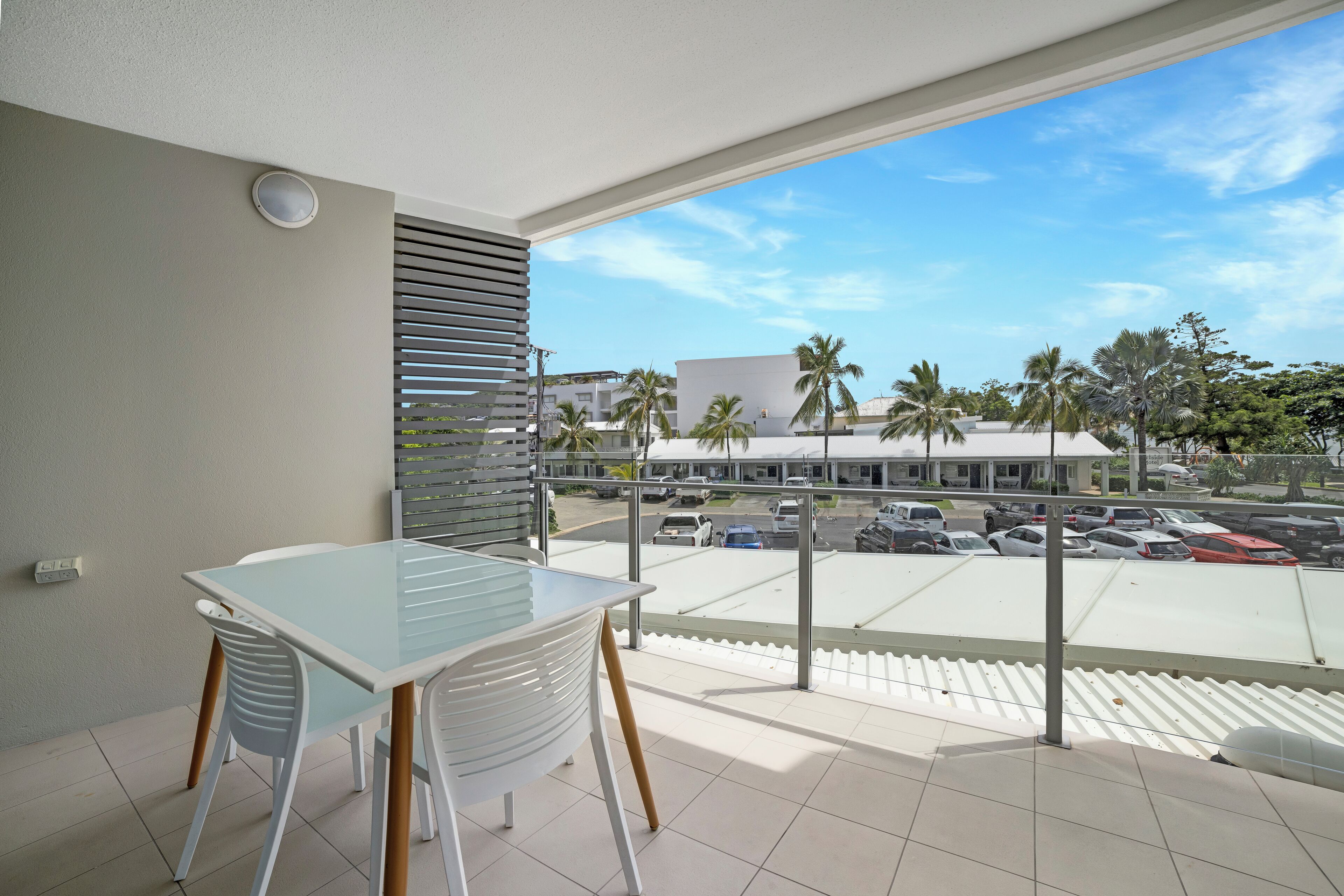 A balcony with a glass table and chairs, overlooking a parking area and buildings with palm trees.