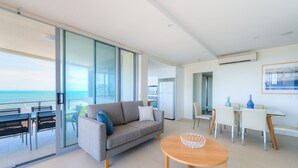 A modern living room with a grey sofa, a round wooden coffee table, and a glass sliding door leading to a balcony with a view of the sea. - Salt Apartments Yeppoon (Yeppoon)