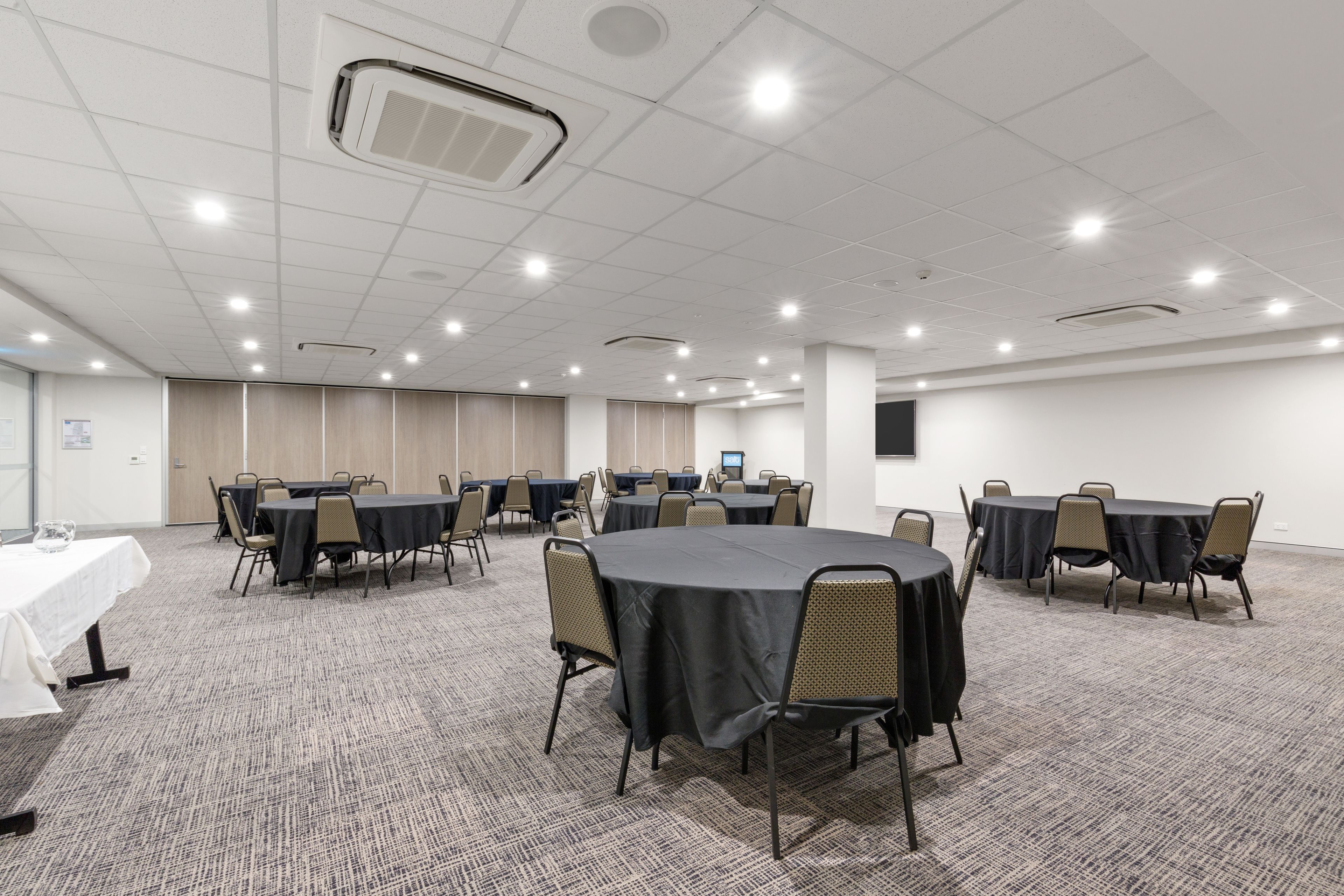 A conference room with multiple tables and chairs, carpeted flooring, and recessed lighting.