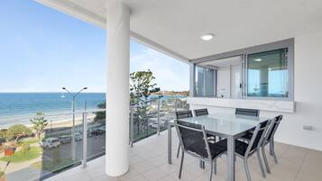 Un balcon avec une table et des chaises en verre, donnant sur une plage et l’océan.