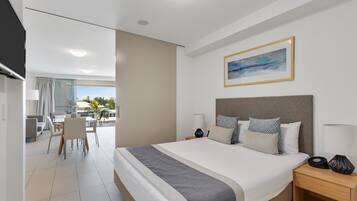 Una habitación de hotel moderna con una cama grande, mesitas de noche, un área de comedor con sillas y vistas al exterior.