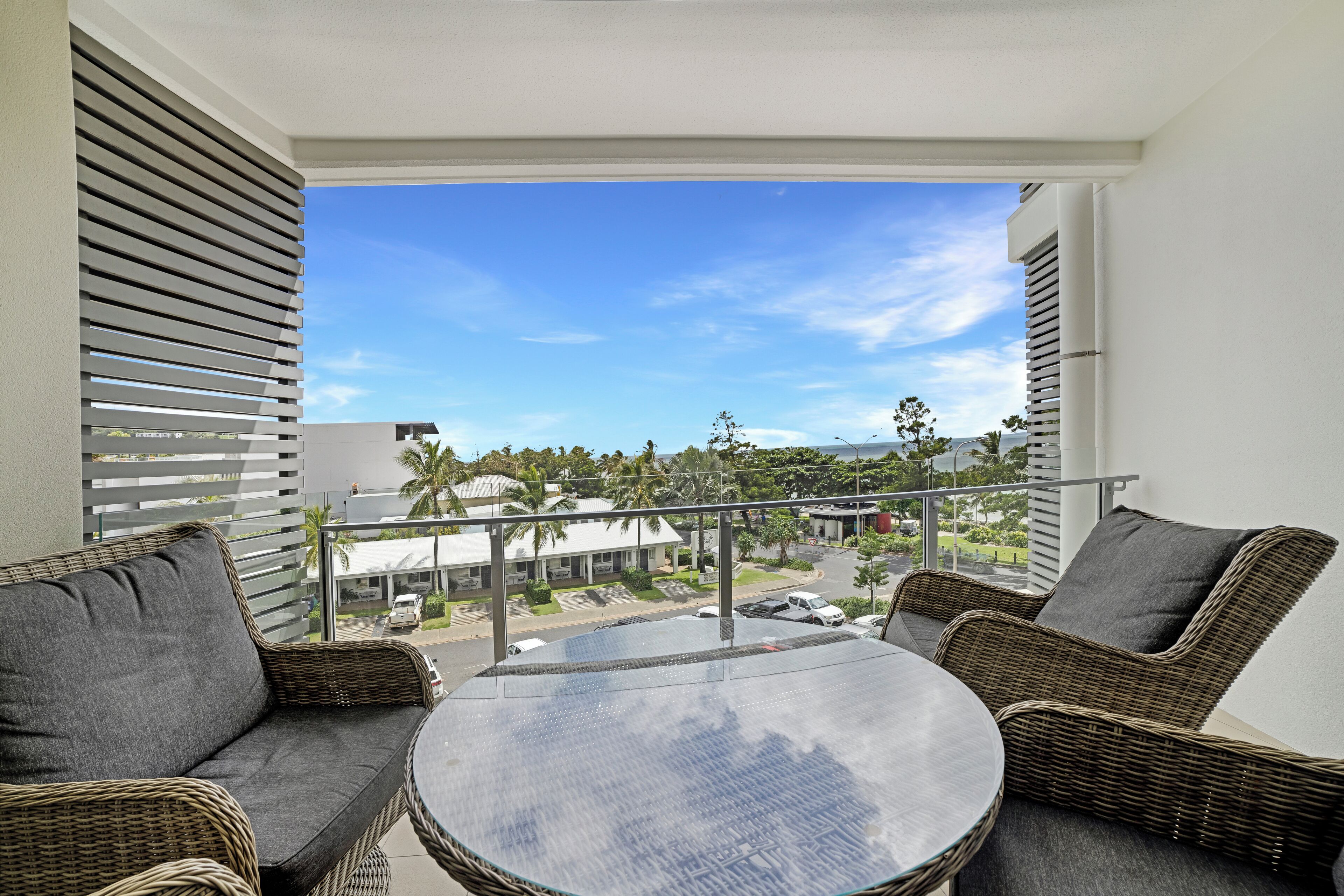 A balcony with wicker furniture and a glass table, overlooking a street with palm trees and buildings.