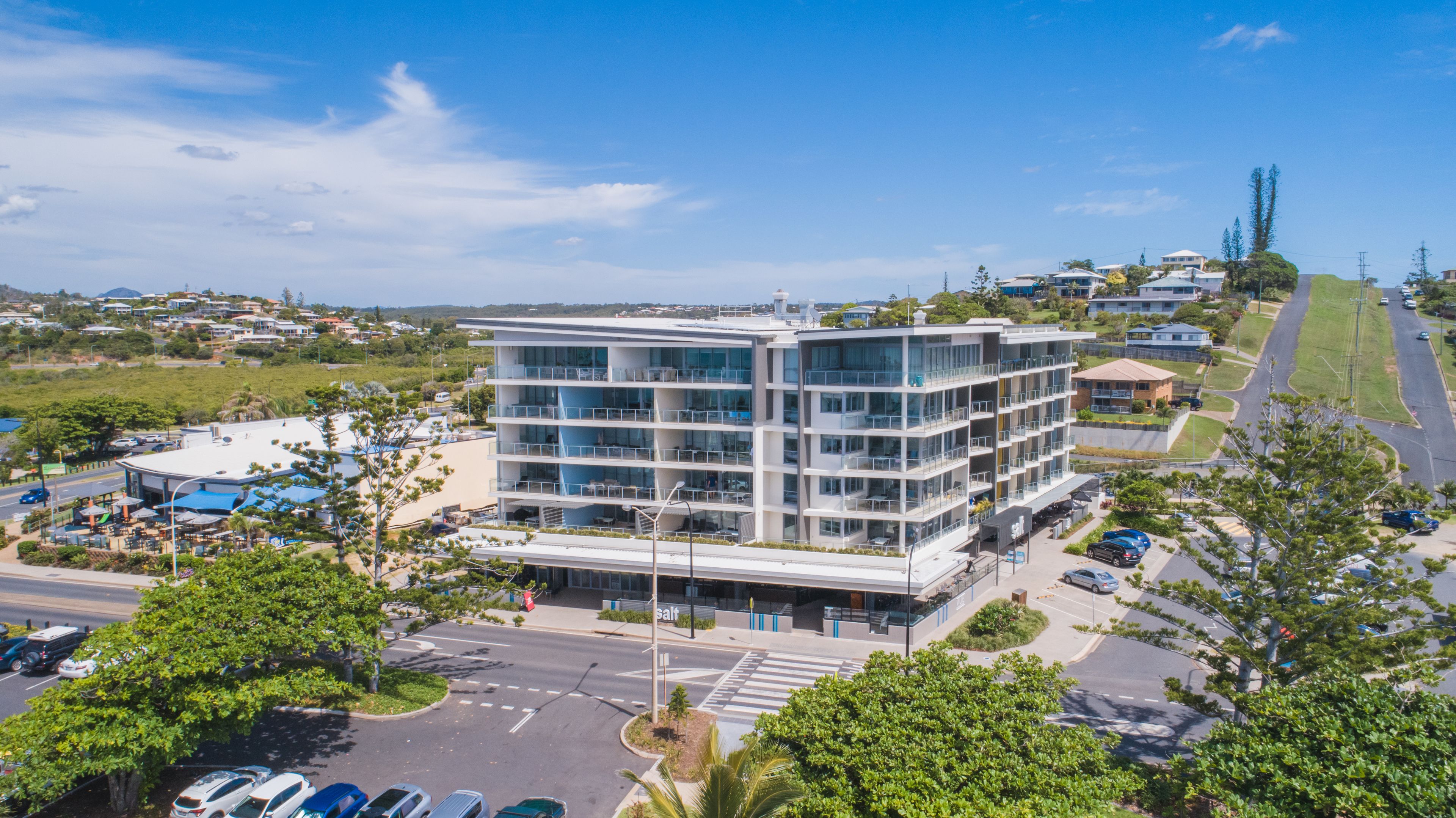 A modern multi-story hotel with a glass facade, located at a street intersection with a parking lot in front.
