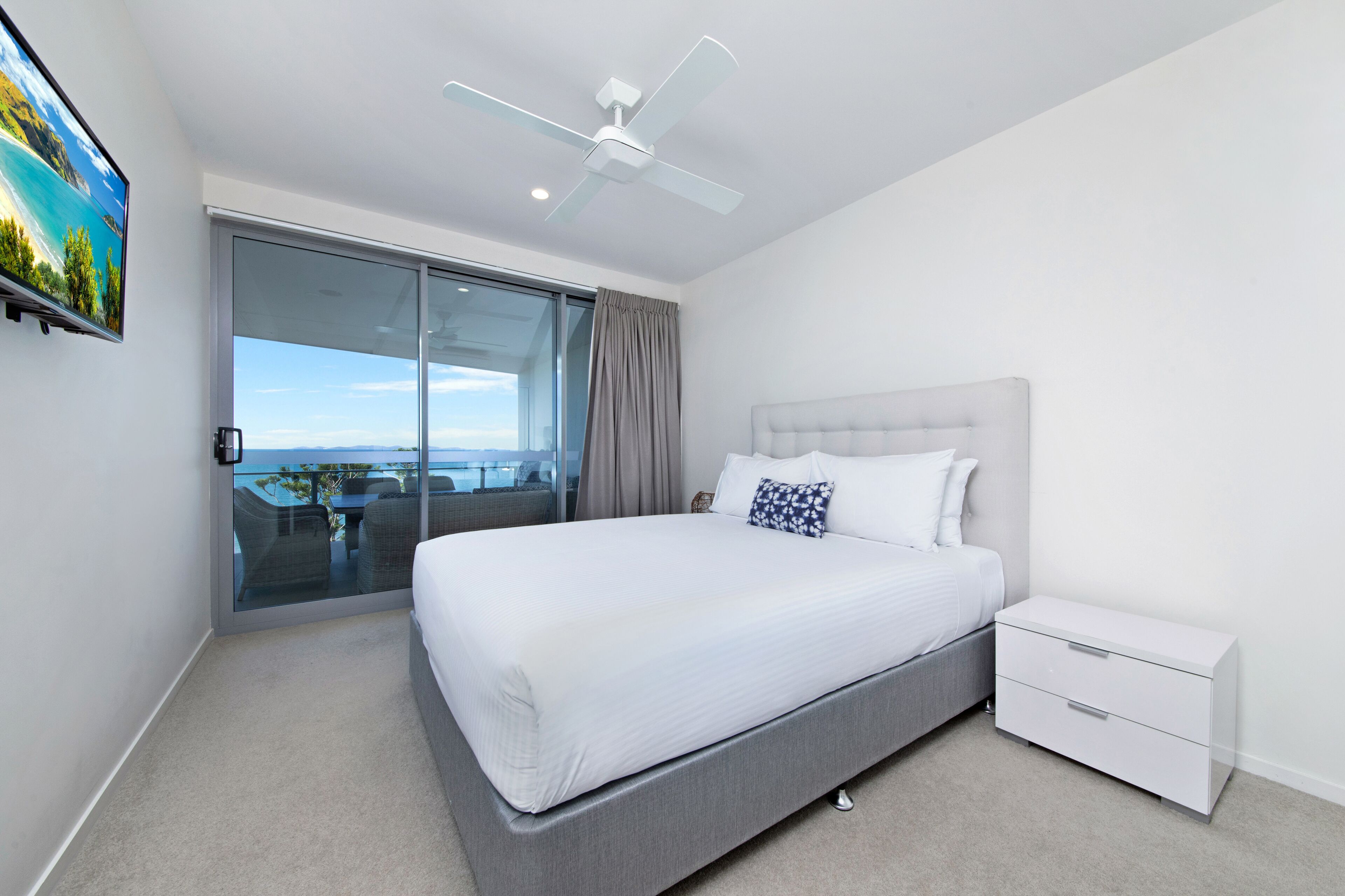 A modern bedroom with a large bed, a nightstand, a ceiling fan, and a sliding glass door leading to a balcony.