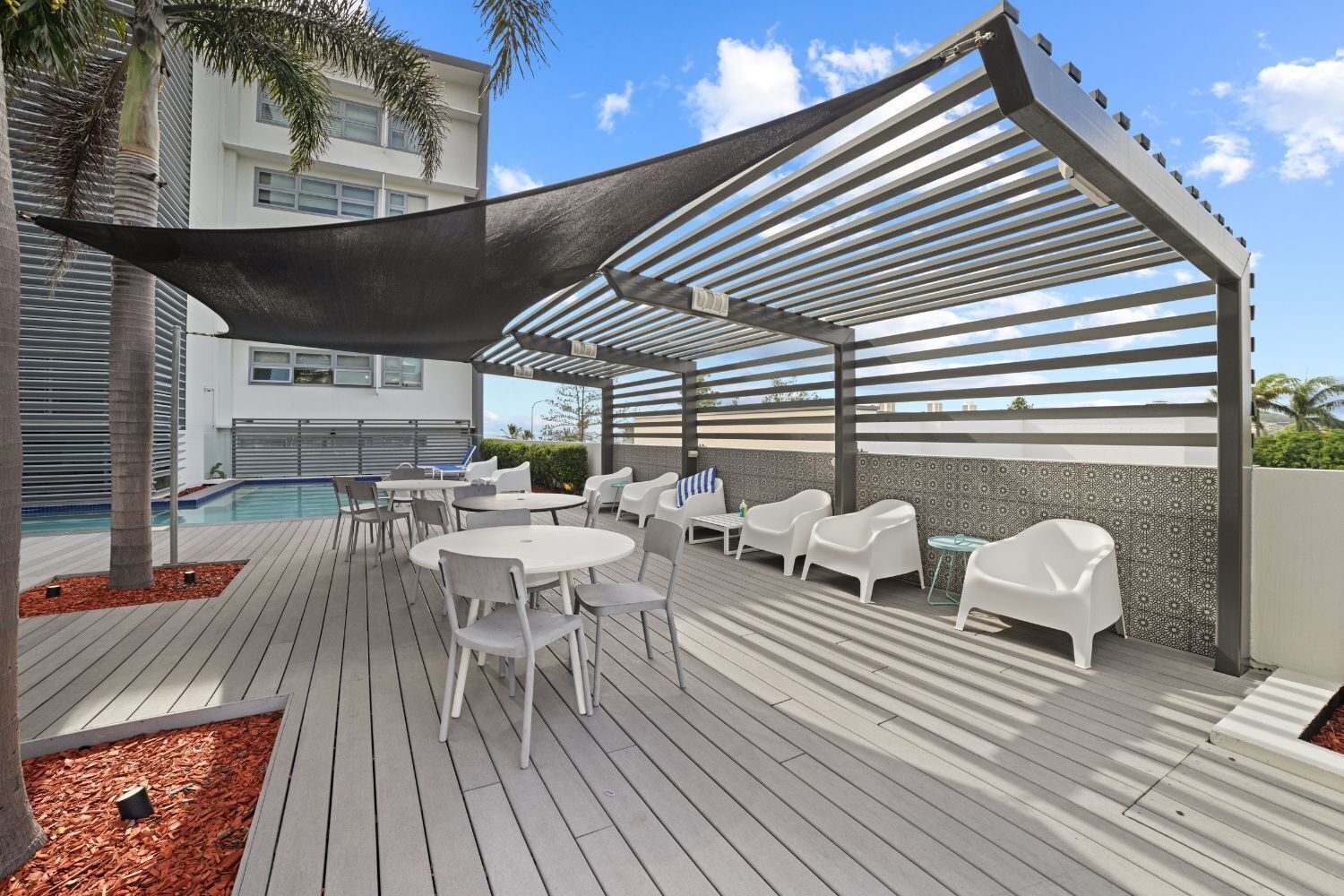 Un espacio al aire libre techado con piscina, mobiliario blanco y una pérgola que ofrece sombra.