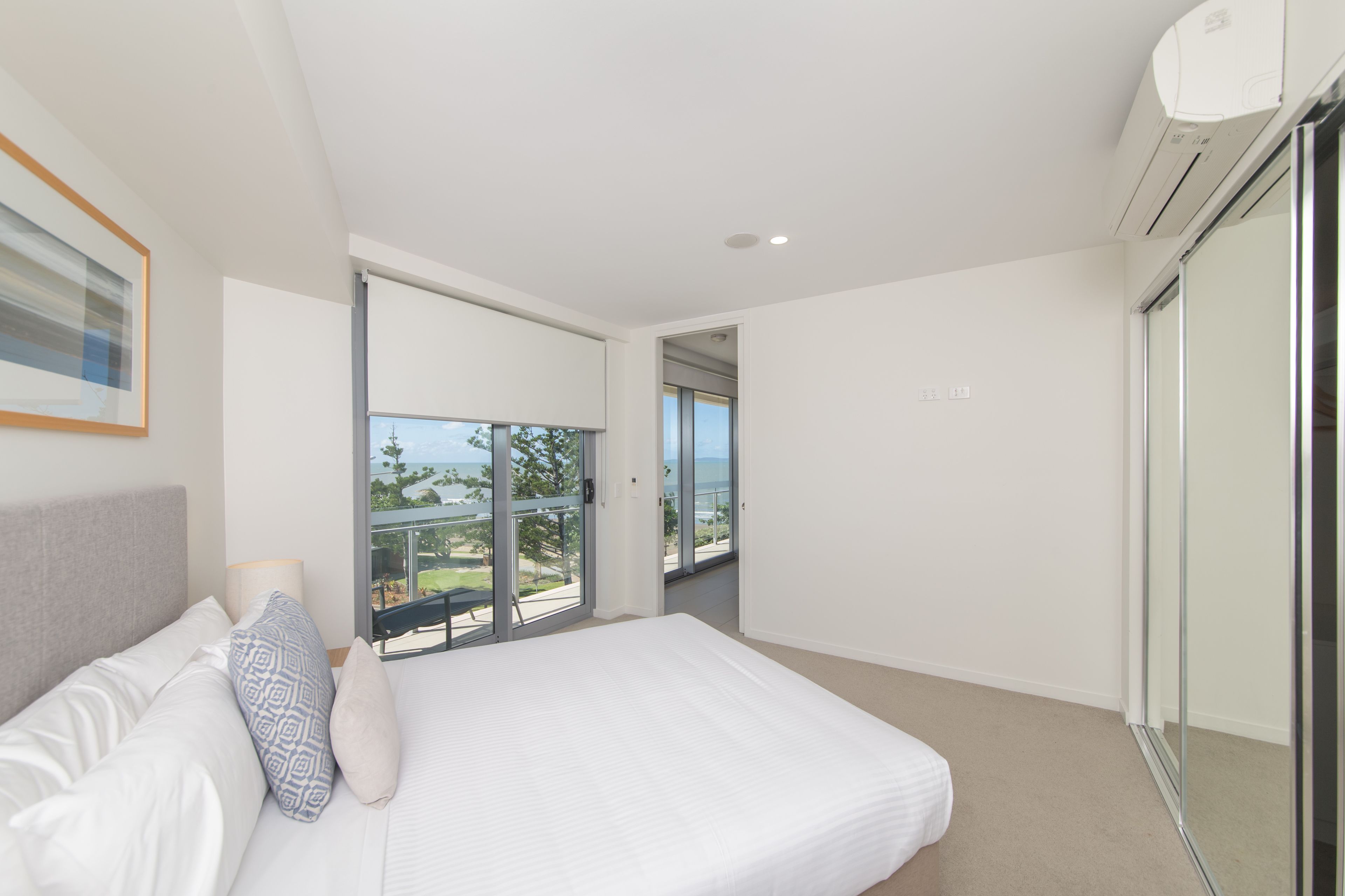 A modern bedroom with a large bed, a balcony with a glass door, and a view of the outdoors.