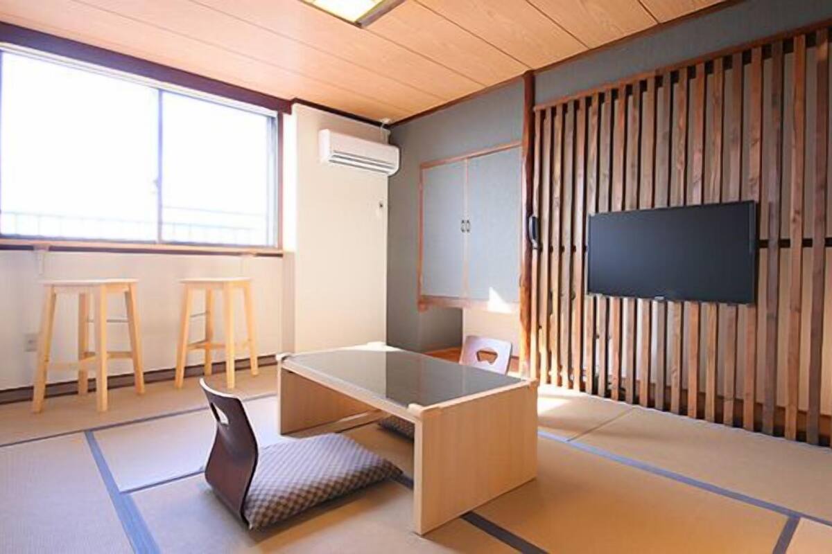 A traditional Japanese room with tatami flooring, a low wooden desk, two stools, a television, and a sliding door.