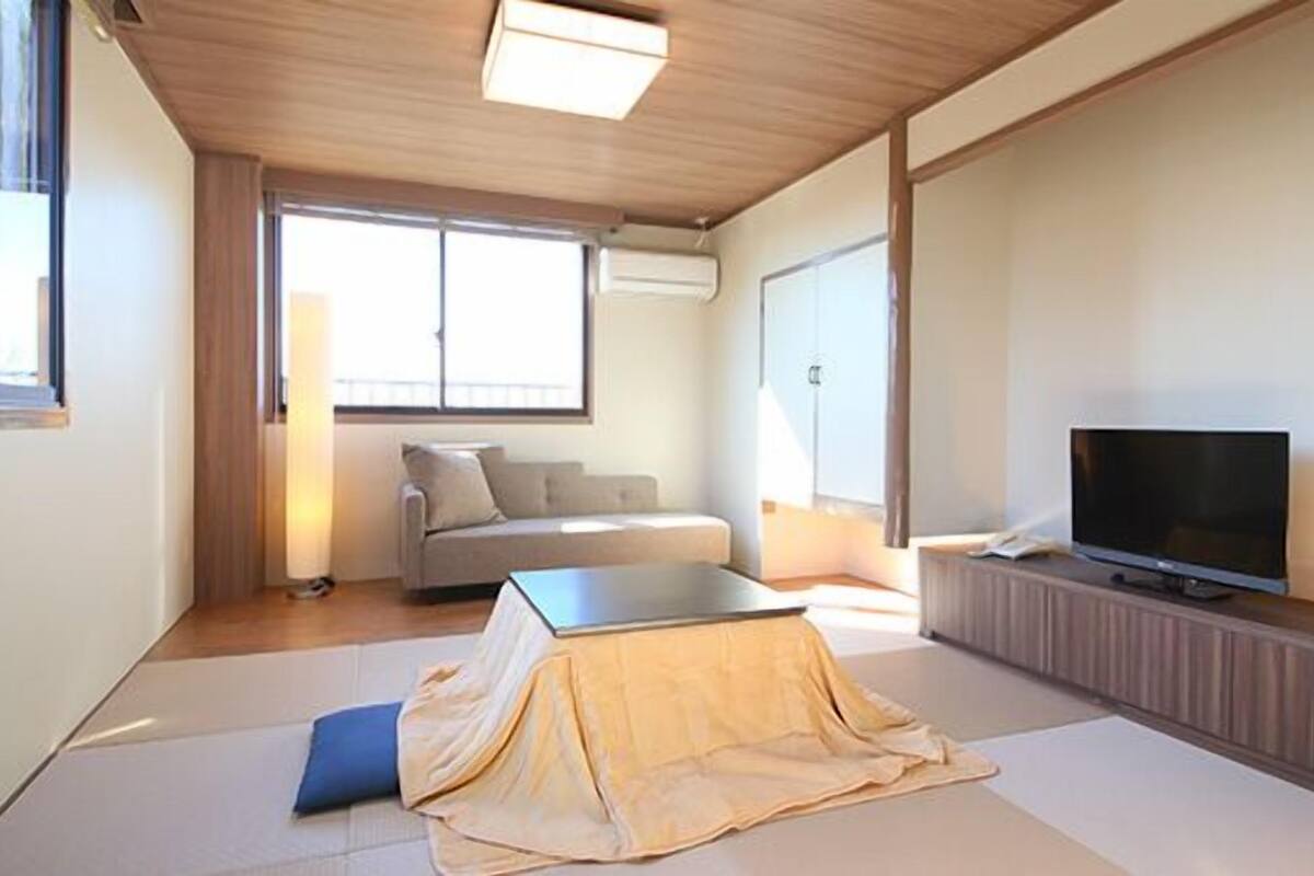 A modern, minimalist living room with tatami flooring, a sofa, a television, and a large window providing natural light.