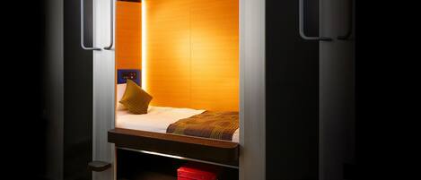 A compact room with a bed, a red suitcase, and a control panel on the wall.
