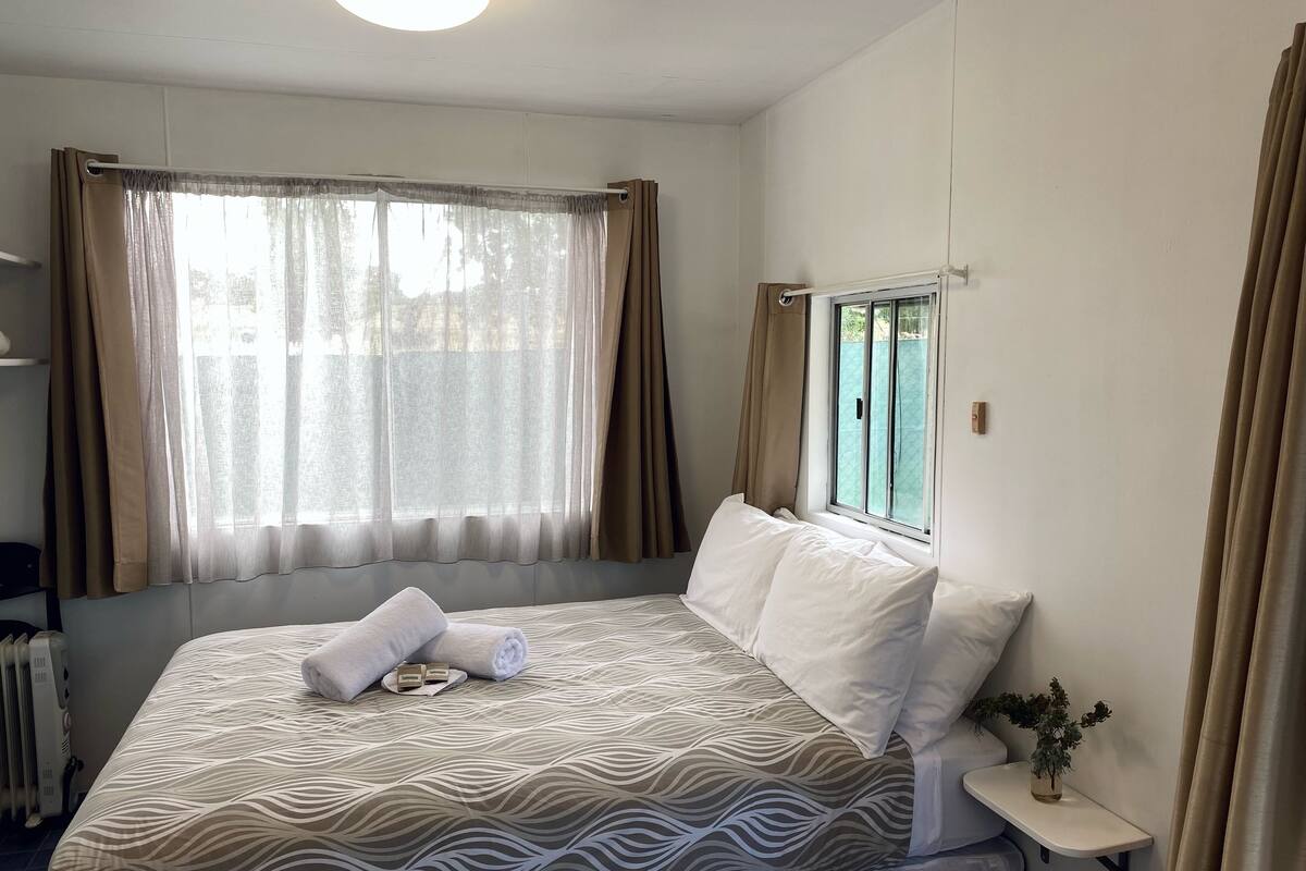 A neatly made bed with a patterned bedspread, a bedside table with a plant, and a window with curtains.