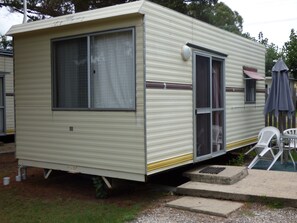 A mobile home with a large window, a sliding door, and a small porch area with a chair and umbrella. - Tenterfield Lodge and Caravan Park (Tenterfield)