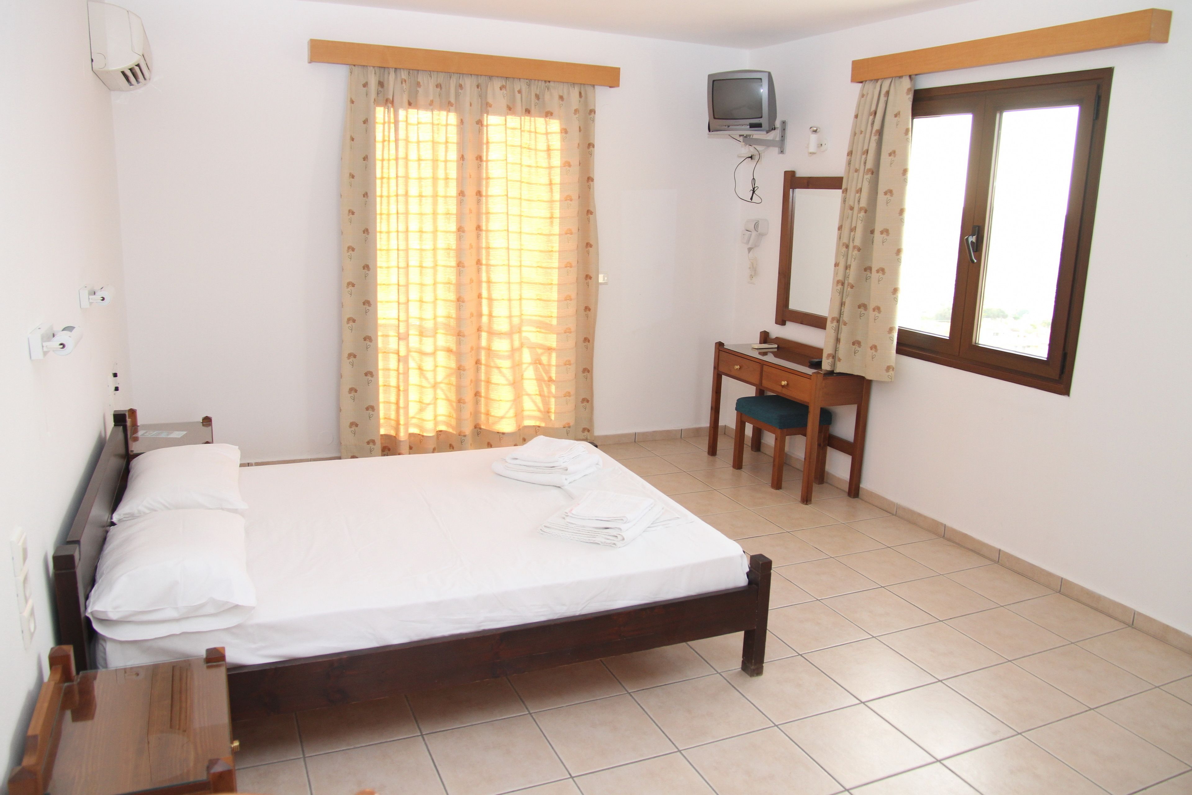 Une chambre à coucher avec un lit, un bureau, une chaise, une télévision et une fenêtre avec des rideaux.