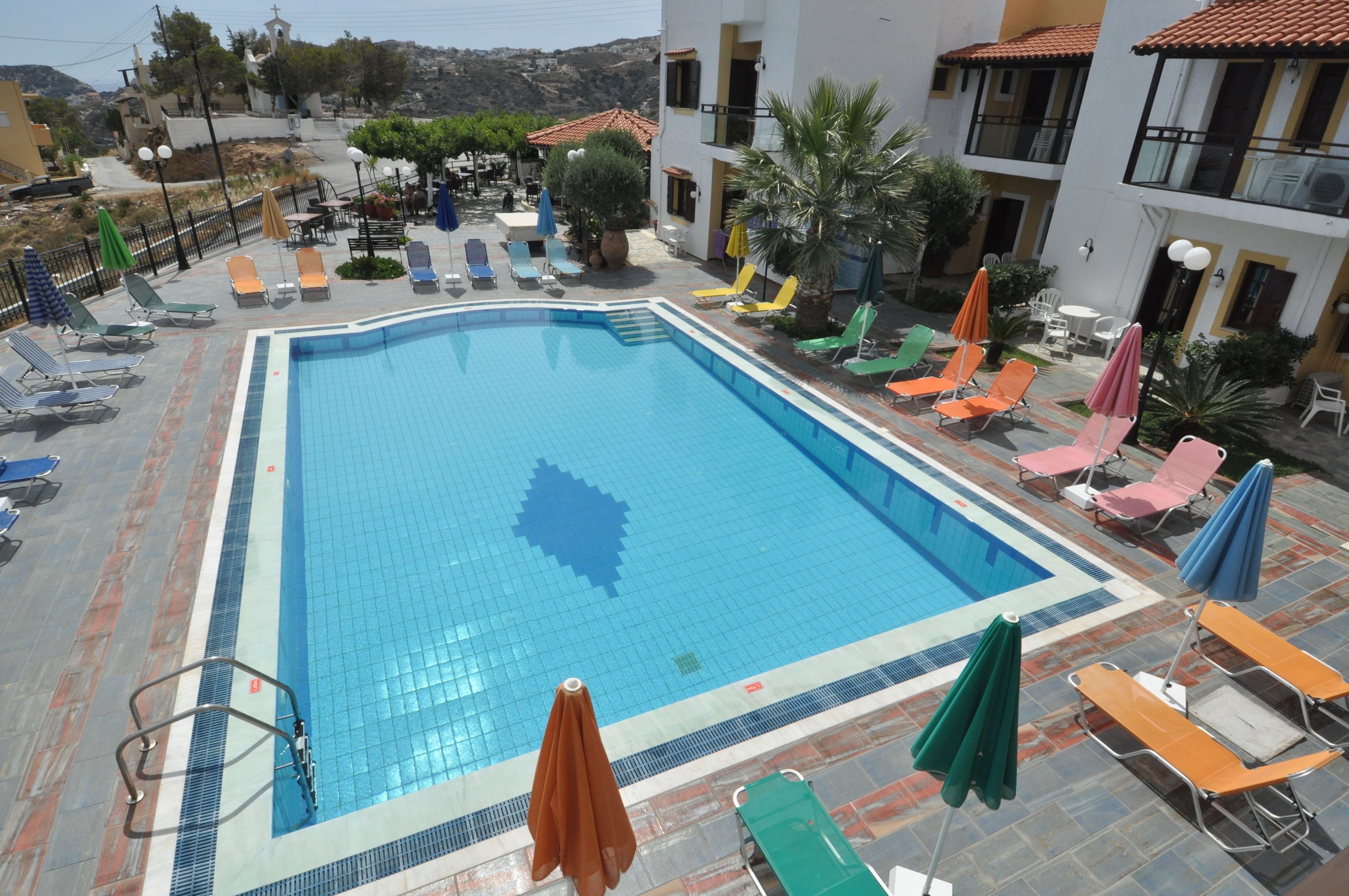 Ein Schwimmbecken mit Liegestühlen und Sonnenschirmen, umgeben von Gebäuden und Grünflächen.
