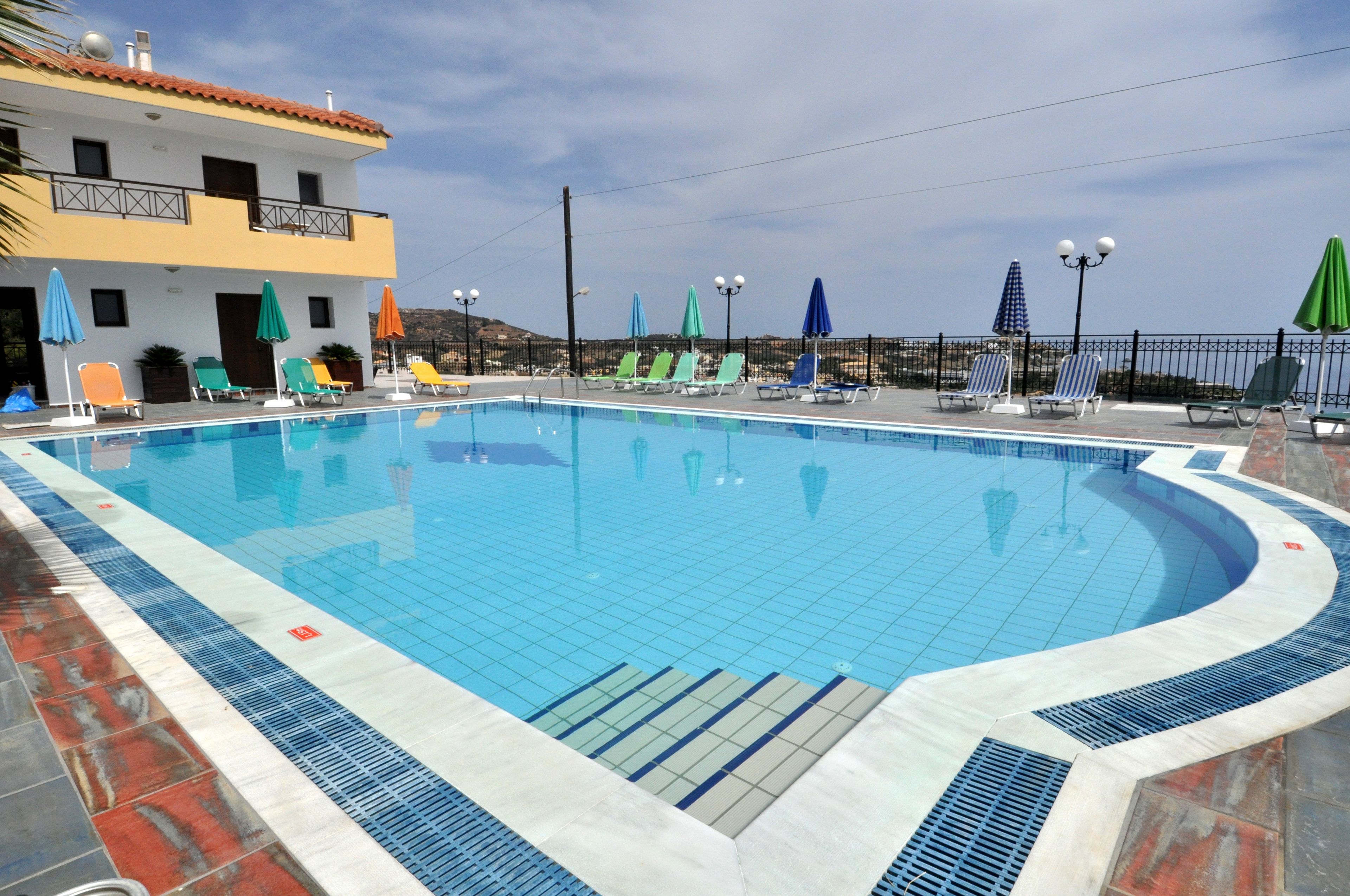 A swimming pool with lounge chairs and umbrellas, surrounded by a building with a tiled roof and a fence.