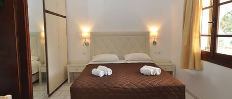 A hotel room with a queen-sized bed, two bedside tables, a mirror, an air conditioning unit, and a window with curtains.
