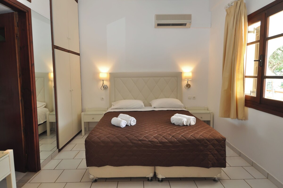 A hotel room with a queen-sized bed, two bedside tables, a mirror, an air conditioning unit, and a window with curtains.